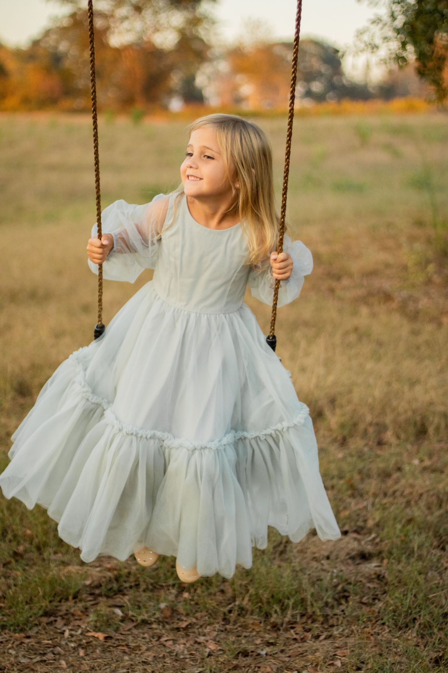 A child in a flowing light blue dress swings at sunset in a rustic outdoor setting.