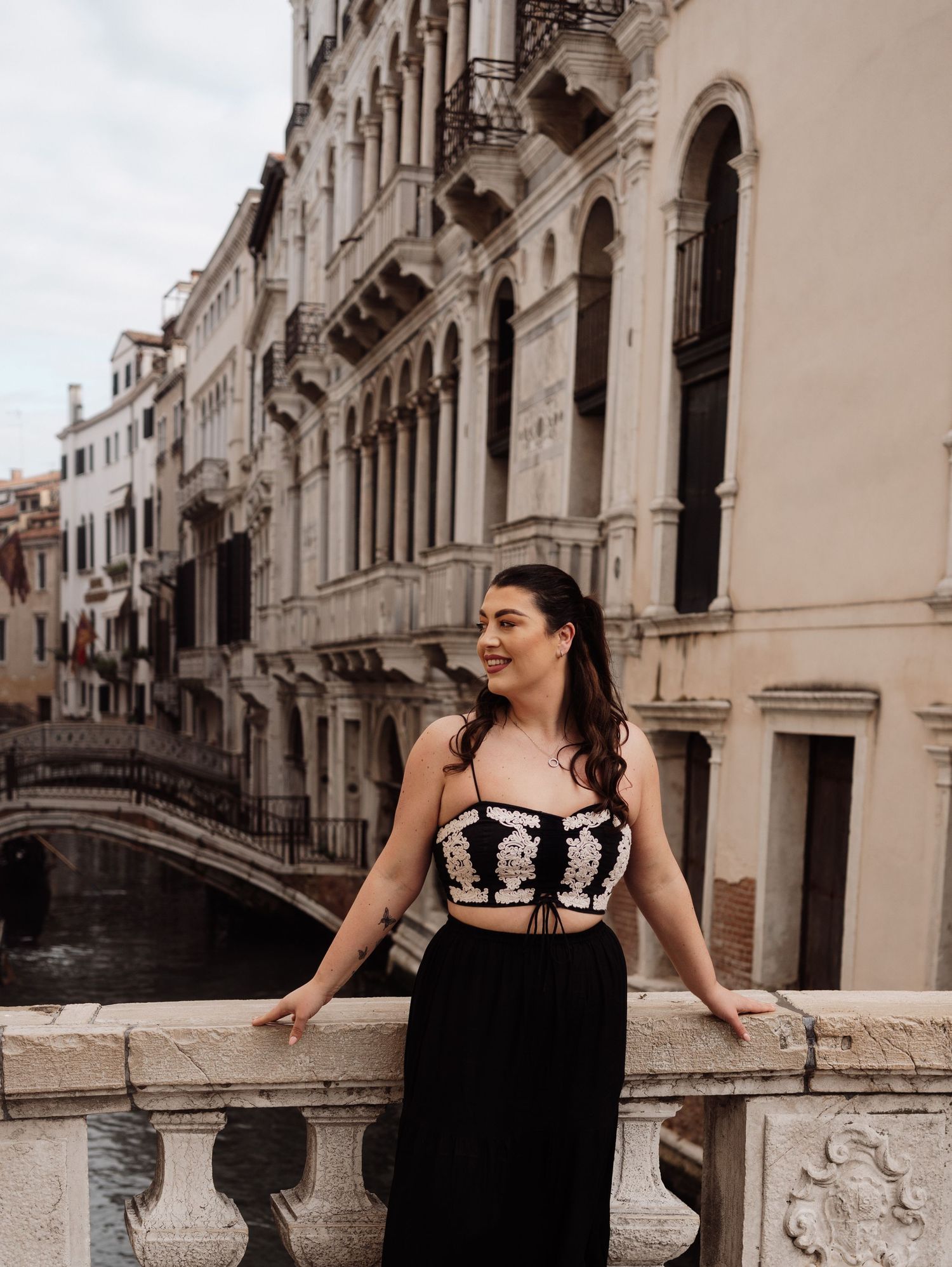 Woman in black and white outfit poses on ornate bridge in Venice with historic buildings and canal in background.