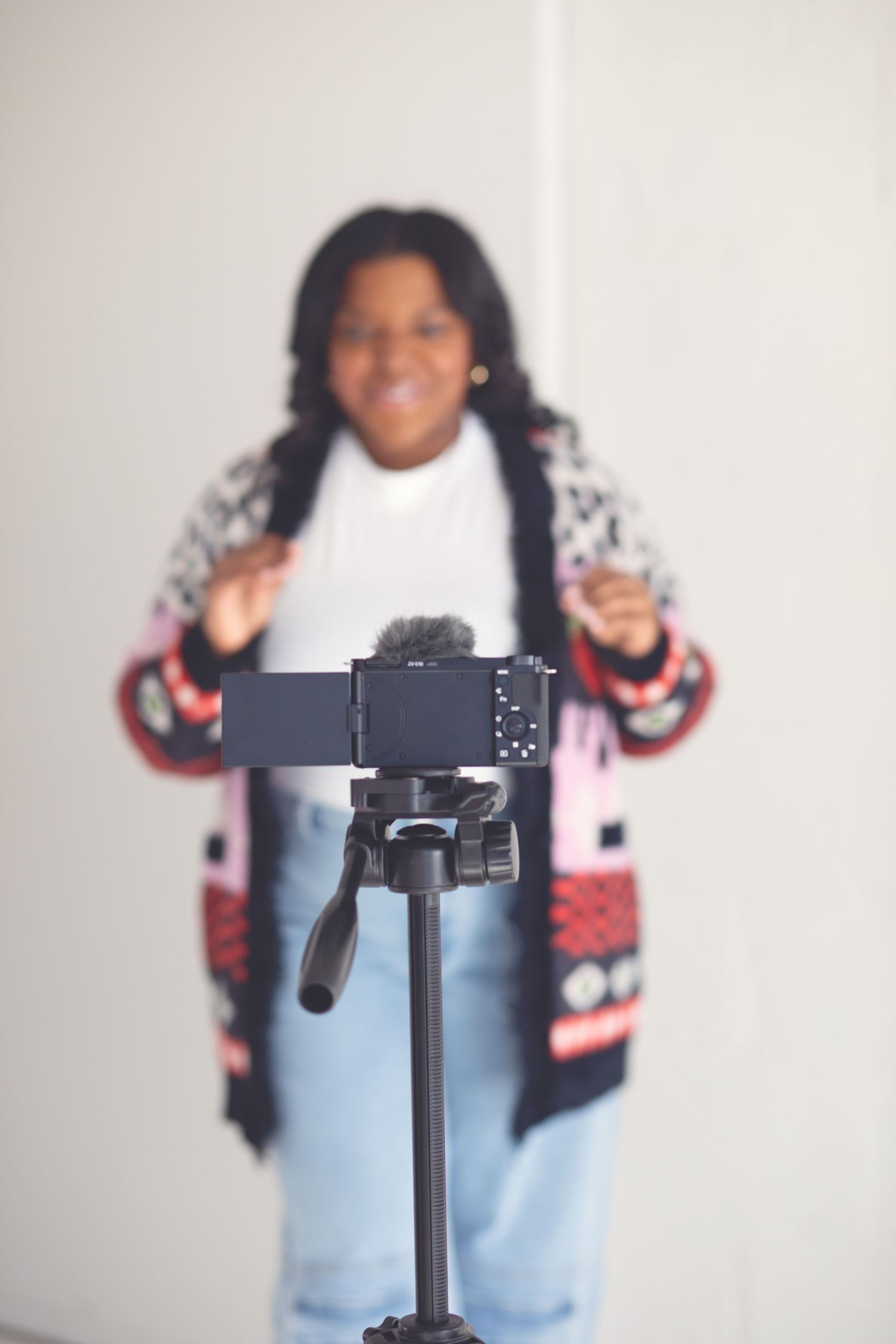 Content creator filming with camera mounted on tripod wearing patterned cardigan and jeans.