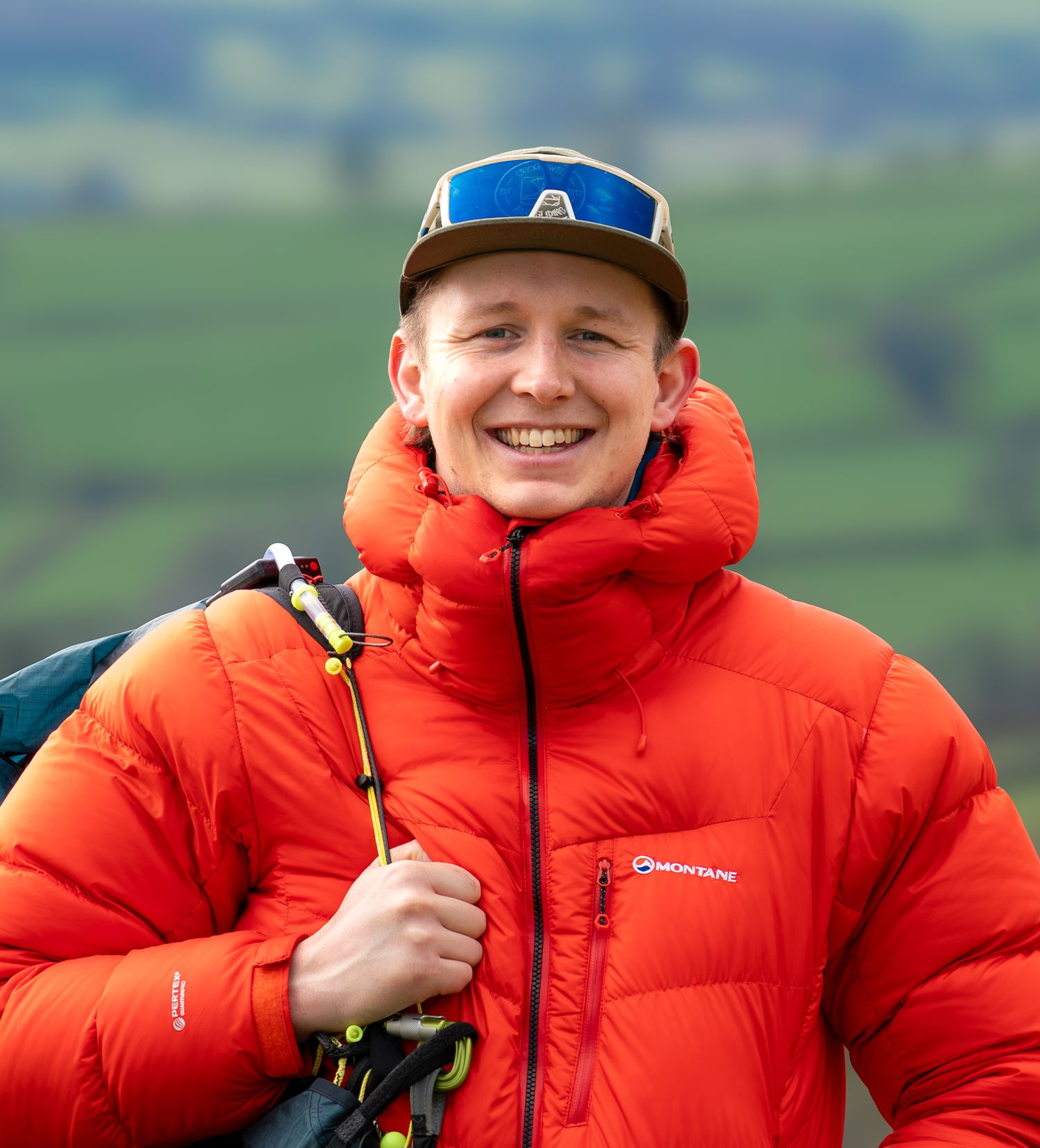 Outdoor adventurer wearing bright orange puffy jacket with baseball cap against green rolling hills background.