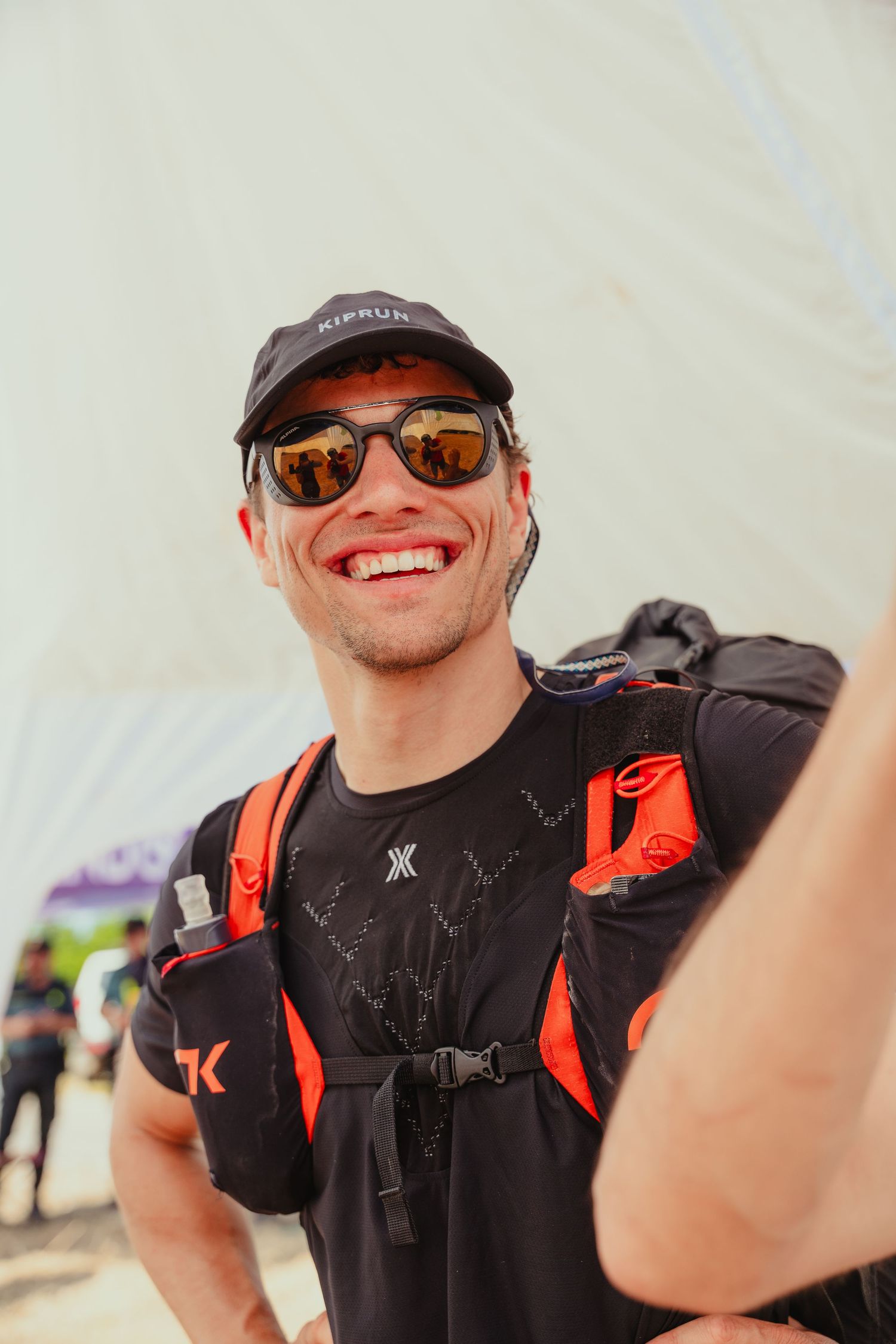 Smiling male trail runner wearing sunglasses, black cap, and orange running vest backpack outdoors.