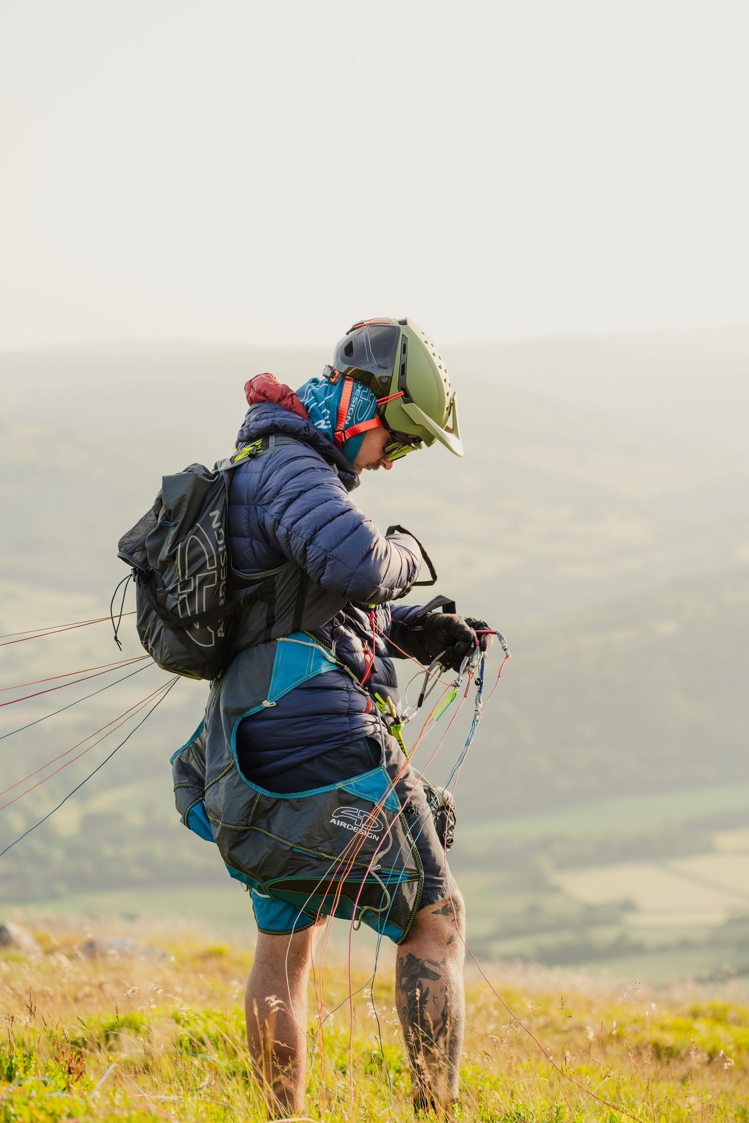 A paraglider prepares for takeoff on a grassy hillside overlooking a scenic valley on a cloudy day.