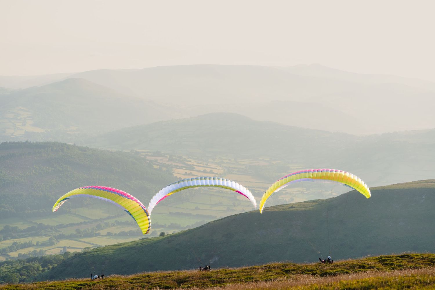 Three colorful paragliders soar above mountainous terrain with misty hills in the background during sunset.