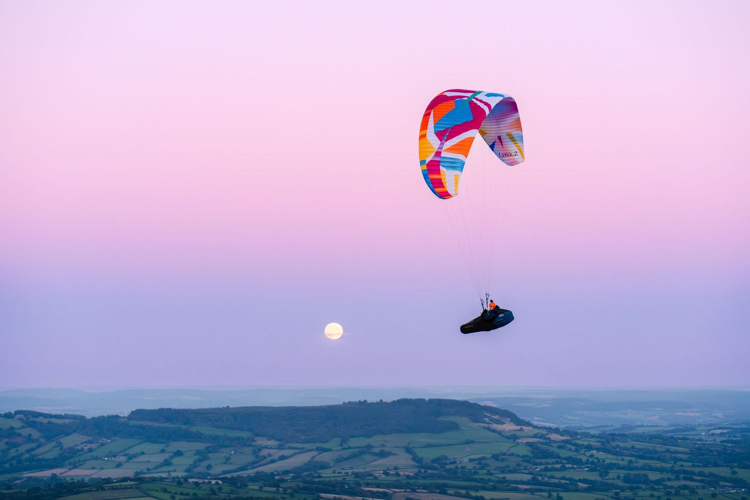 Colorful paraglider soaring over green hills with a full moon rising against a pink and purple twilight sky.