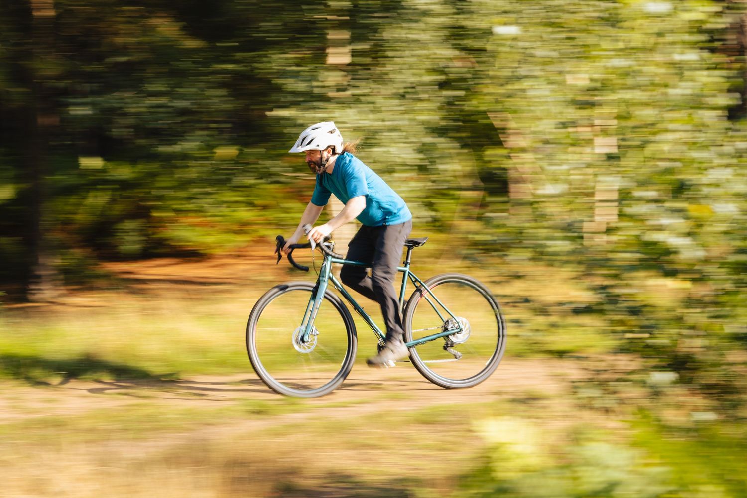 Motion blurred cyclist in blue jersey riding a road bike on a trail with green foliage in the background.