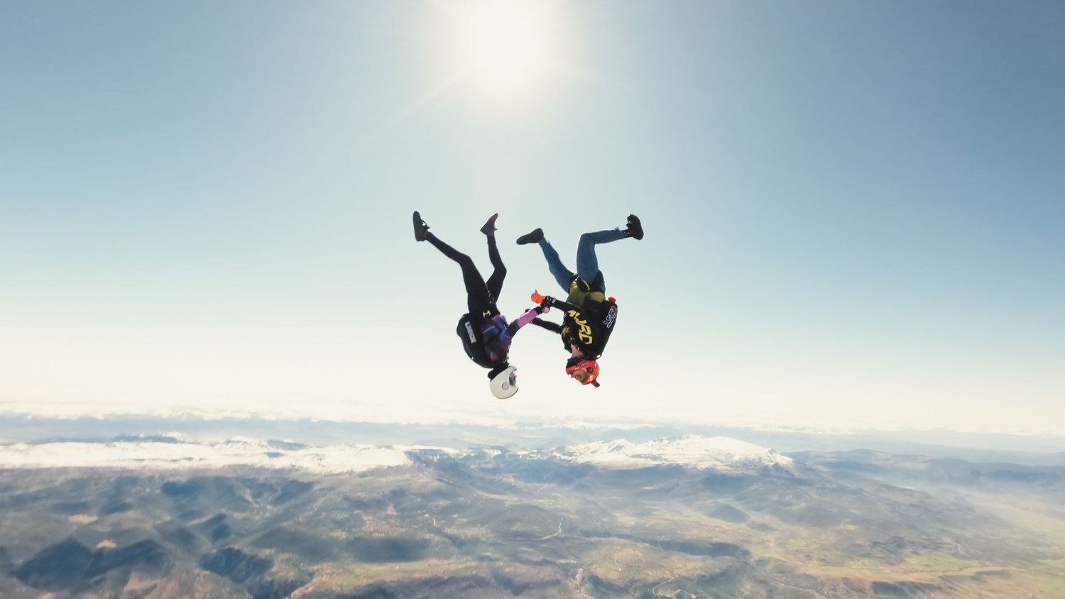 Two skydivers performing headdown formation skydiving high above scenic mountain landscape on a clear sunny day.