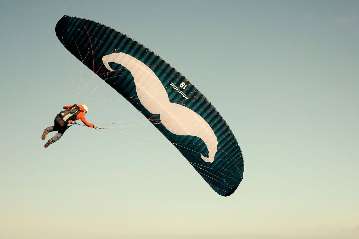 A skydiver descends beneath a dark paragliding wing against a pale sky at sunset.