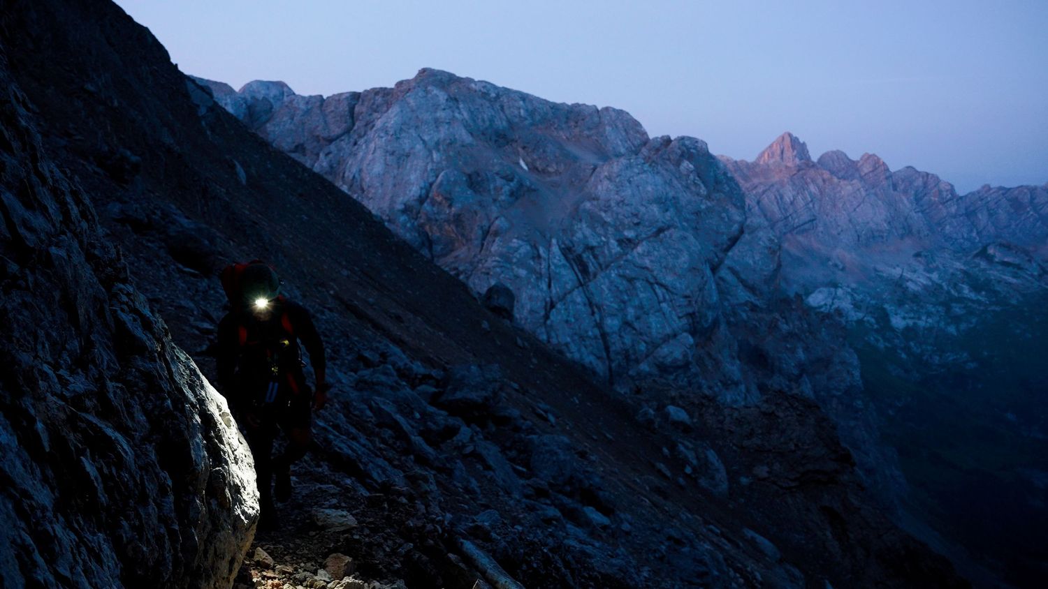 Hiker with headlamp navigating a steep rocky mountain trail at dusk with dramatic peaks in the background.