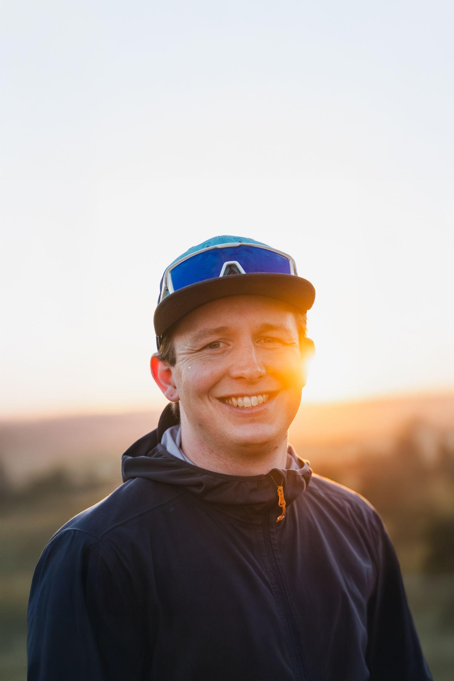 Person wearing a blue baseball cap and black hoodie smiles at sunset in an outdoor setting.