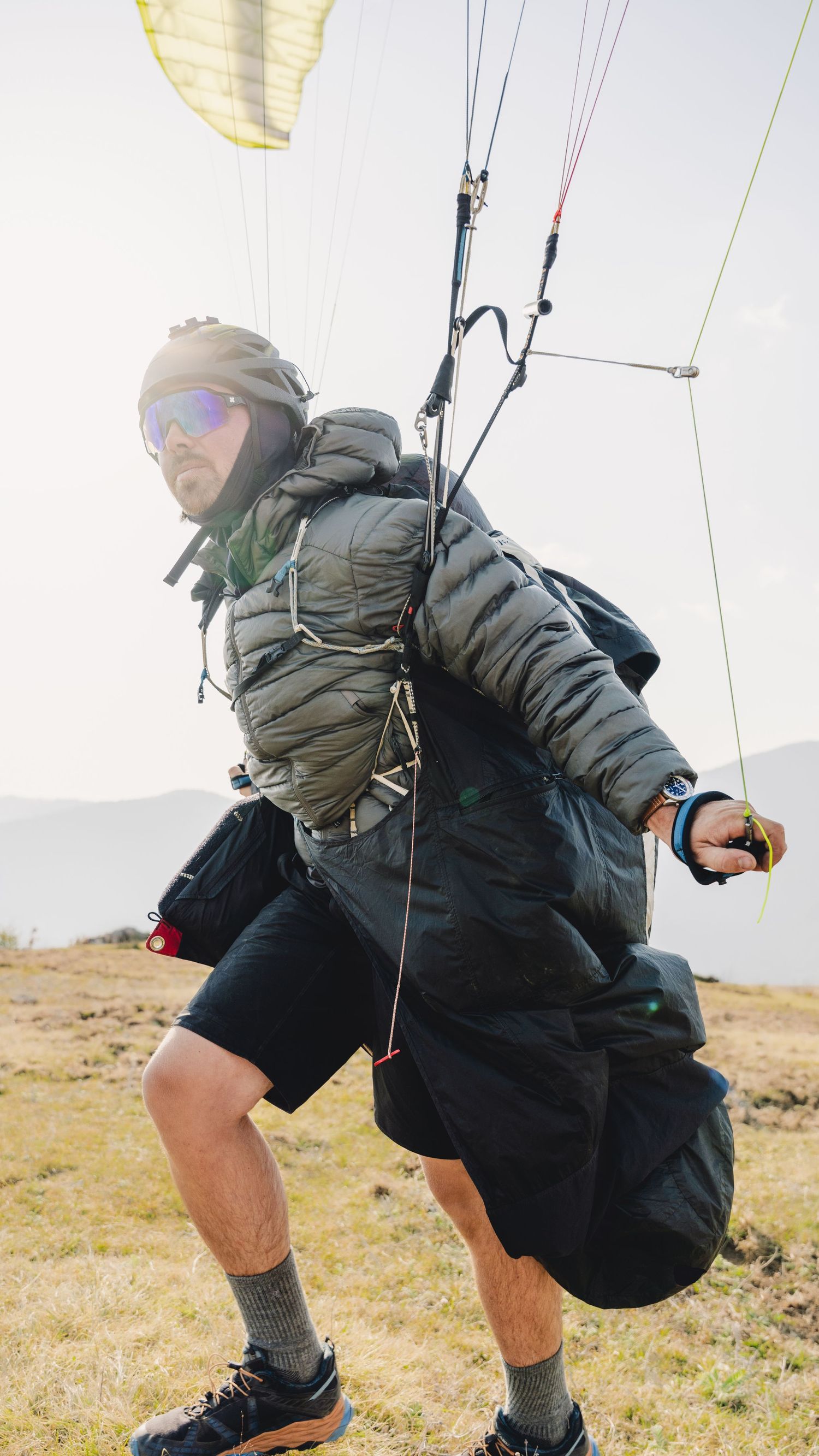 A paraglider in sunglasses and puffer jacket runs on a hillside preparing for takeoff with colorful canopy lines above.