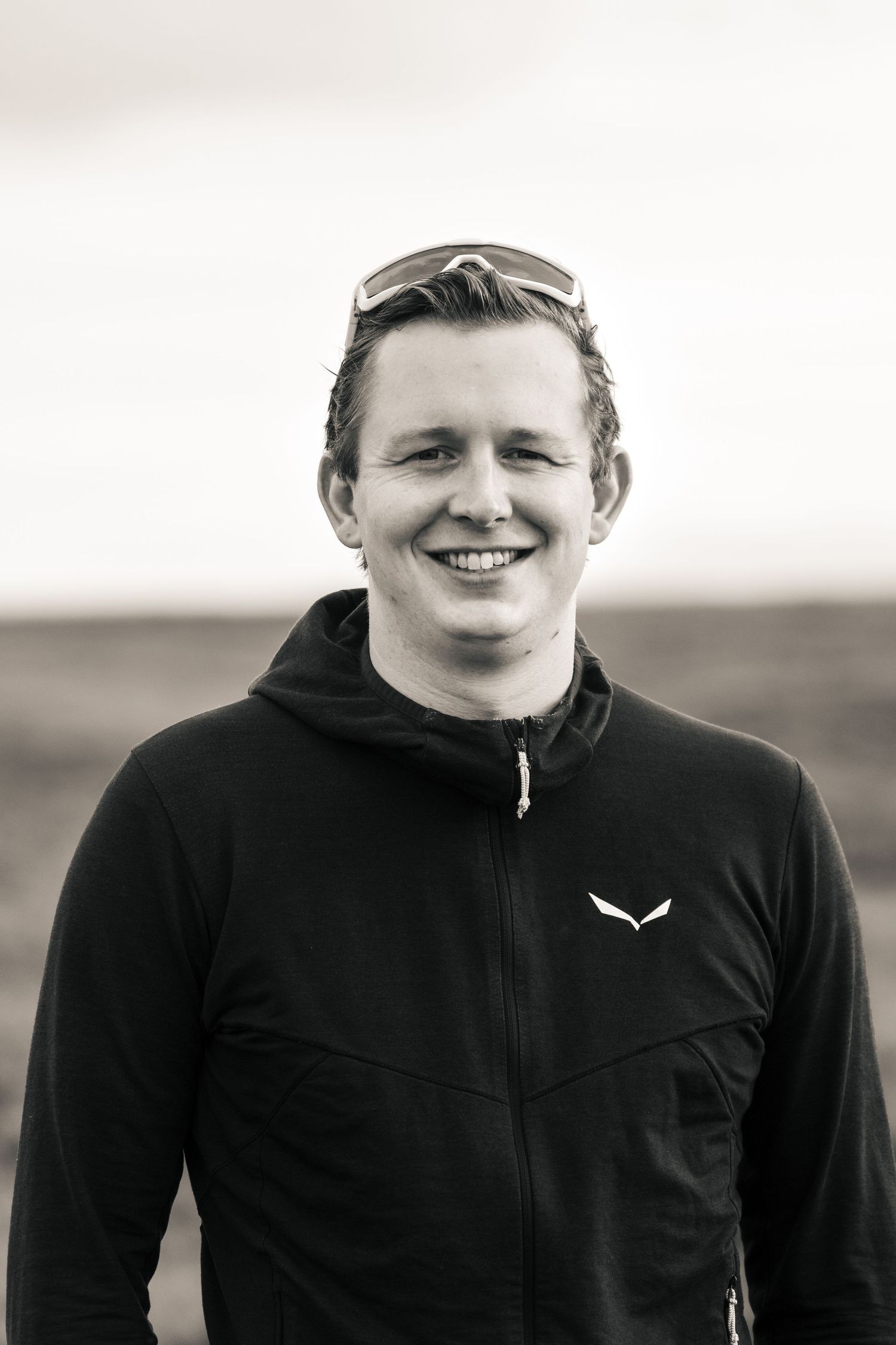 Black and white portrait of a smiling young man wearing a black zip-up jacket with sunglasses on his head outdoors.