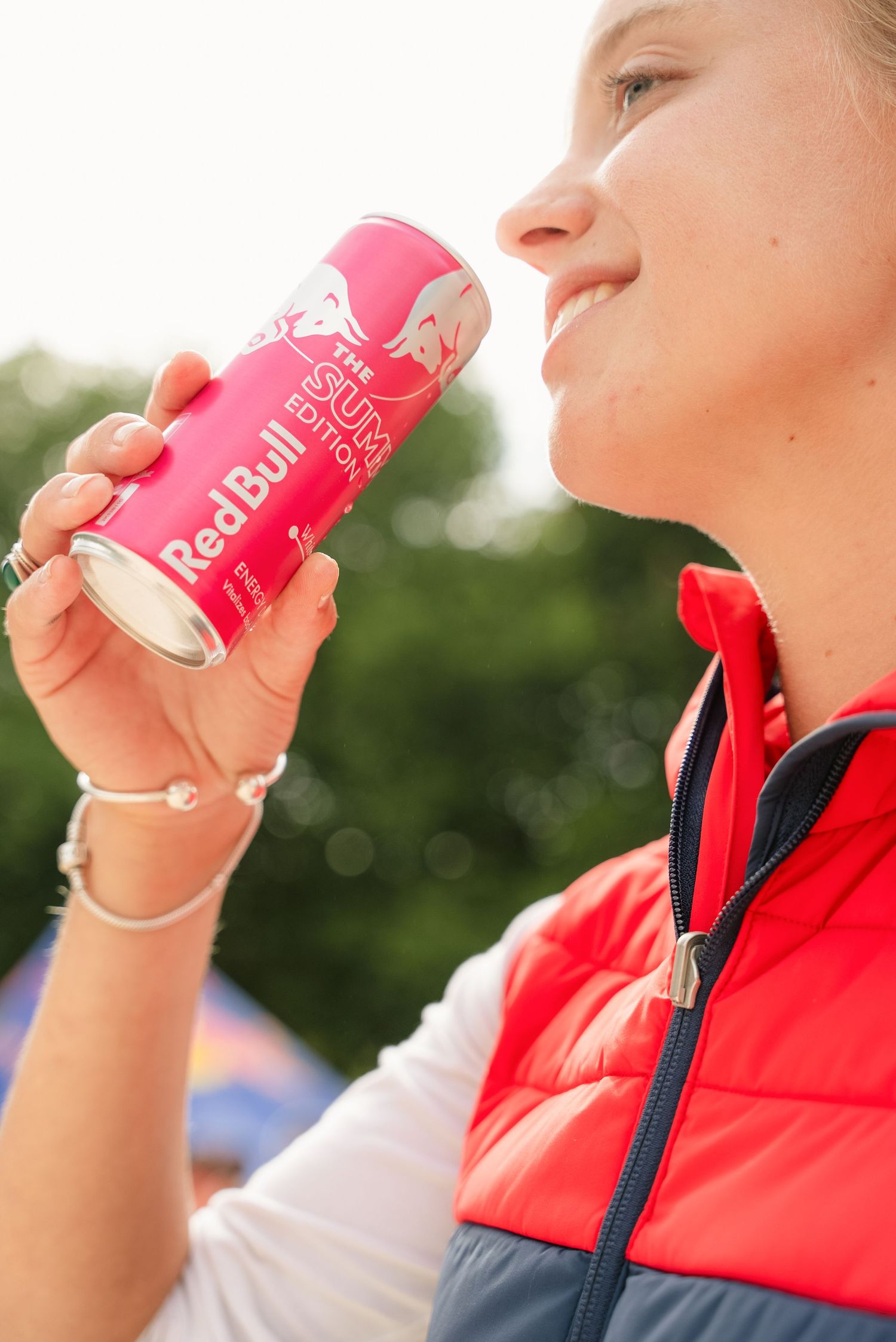 Person in red vest holds and drinks from a pink Red Bull energy drink can outdoors on sunny day.