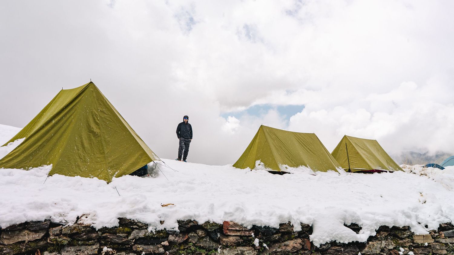 Person standing between yellow tents on snowy mountain campsite with cloudy sky backdrop.