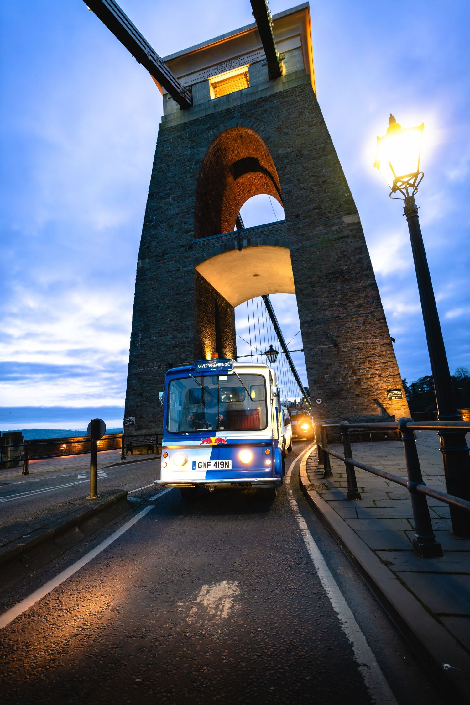 Bus crossing the illuminated Clifton Suspension Bridge at dusk with glowing street lamp and dramatic blue sky.