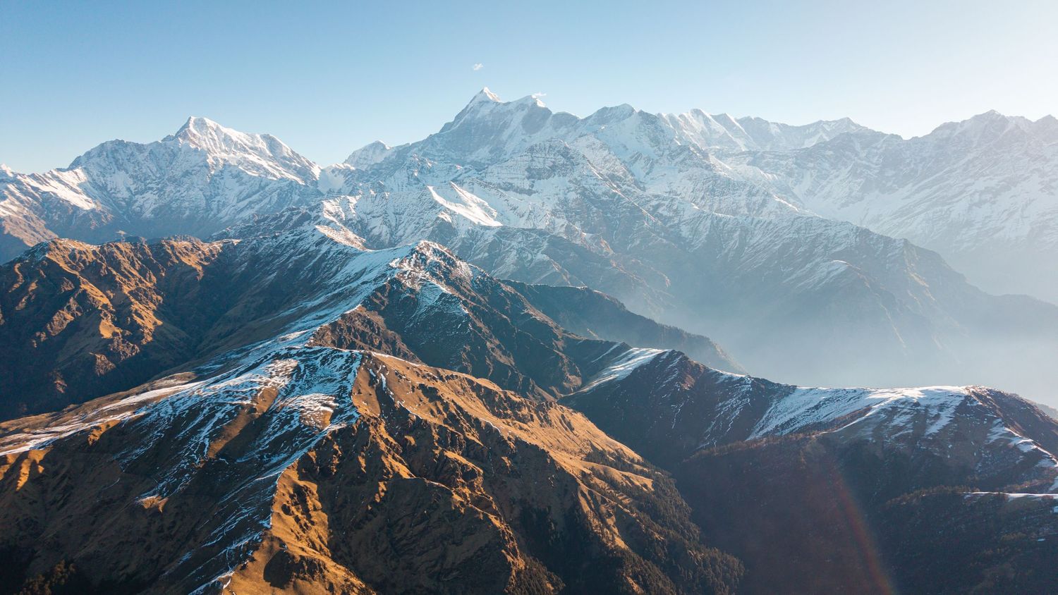 Aerial view of snow-capped Himalayan mountain peaks with golden brown foothills and misty valleys in clear winter light.