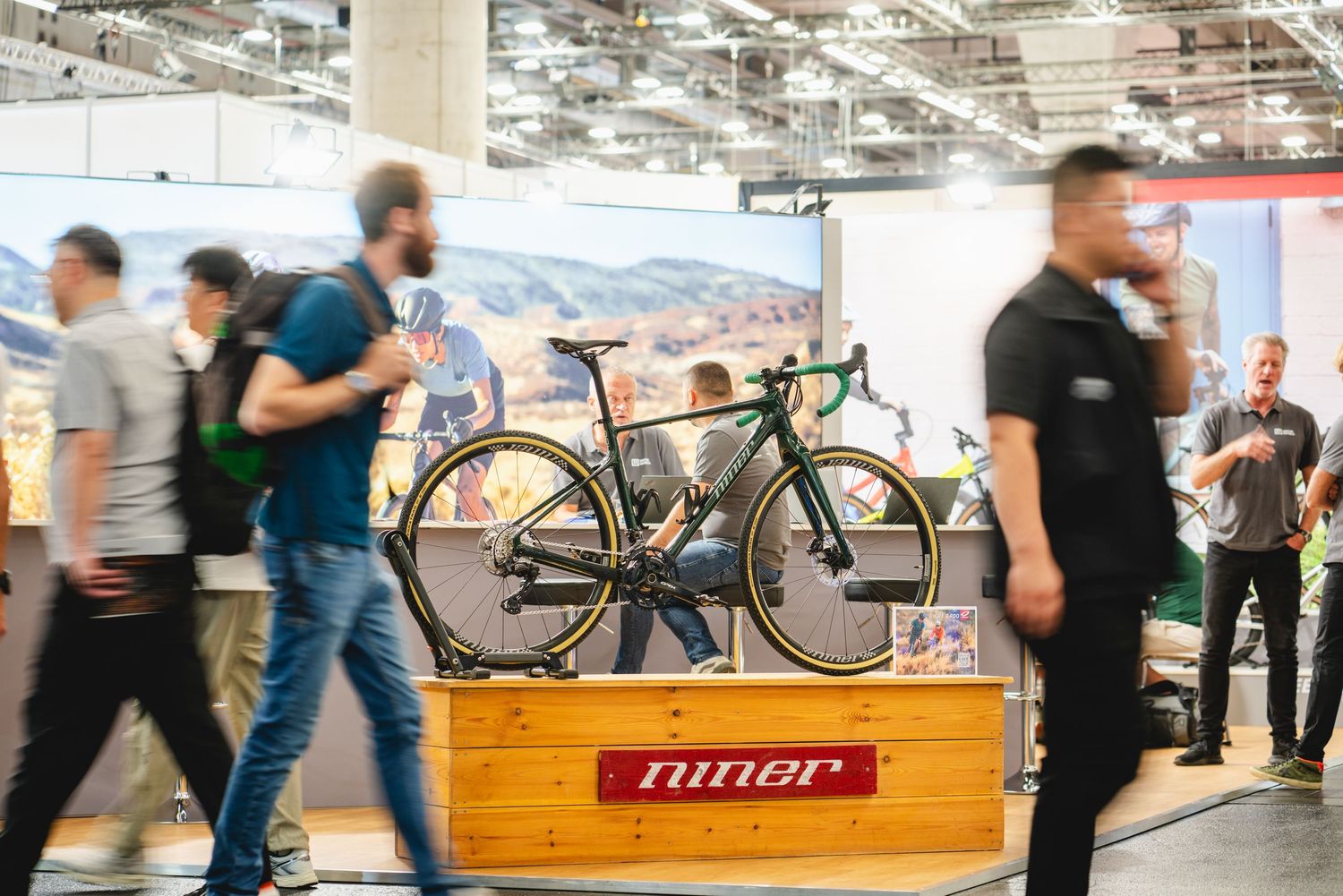 People walking and mingling at an indoor bicycle trade show with displays and wooden platforms visible.
