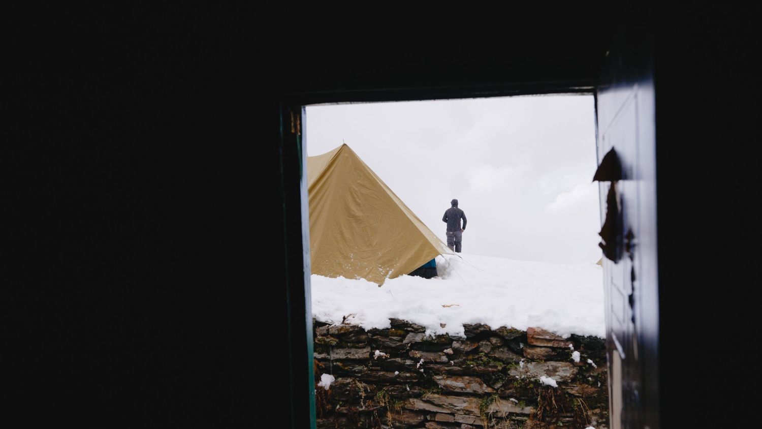 Person standing on snow-covered rubble viewed through dark doorframe with yellow tent visible outside.