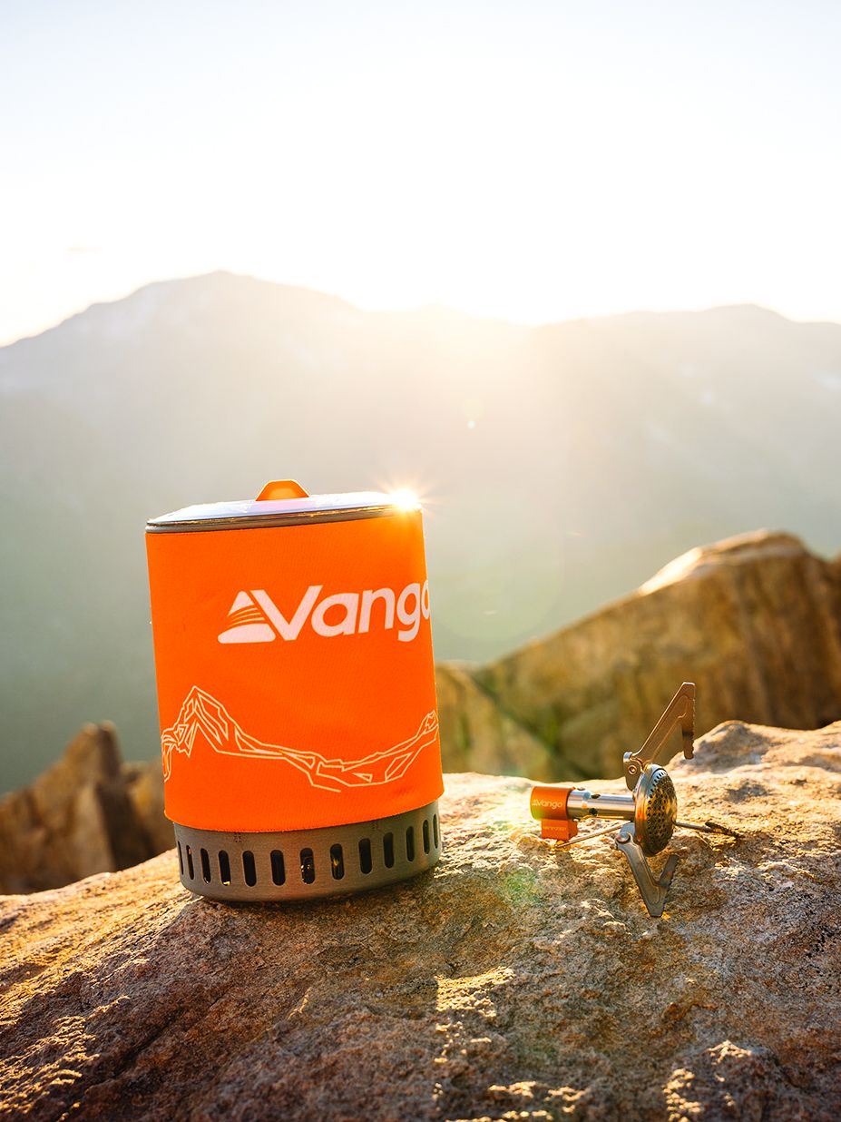 Orange camping stove sits on rocky mountain ledge with scenic misty valley views in background at sunset.
