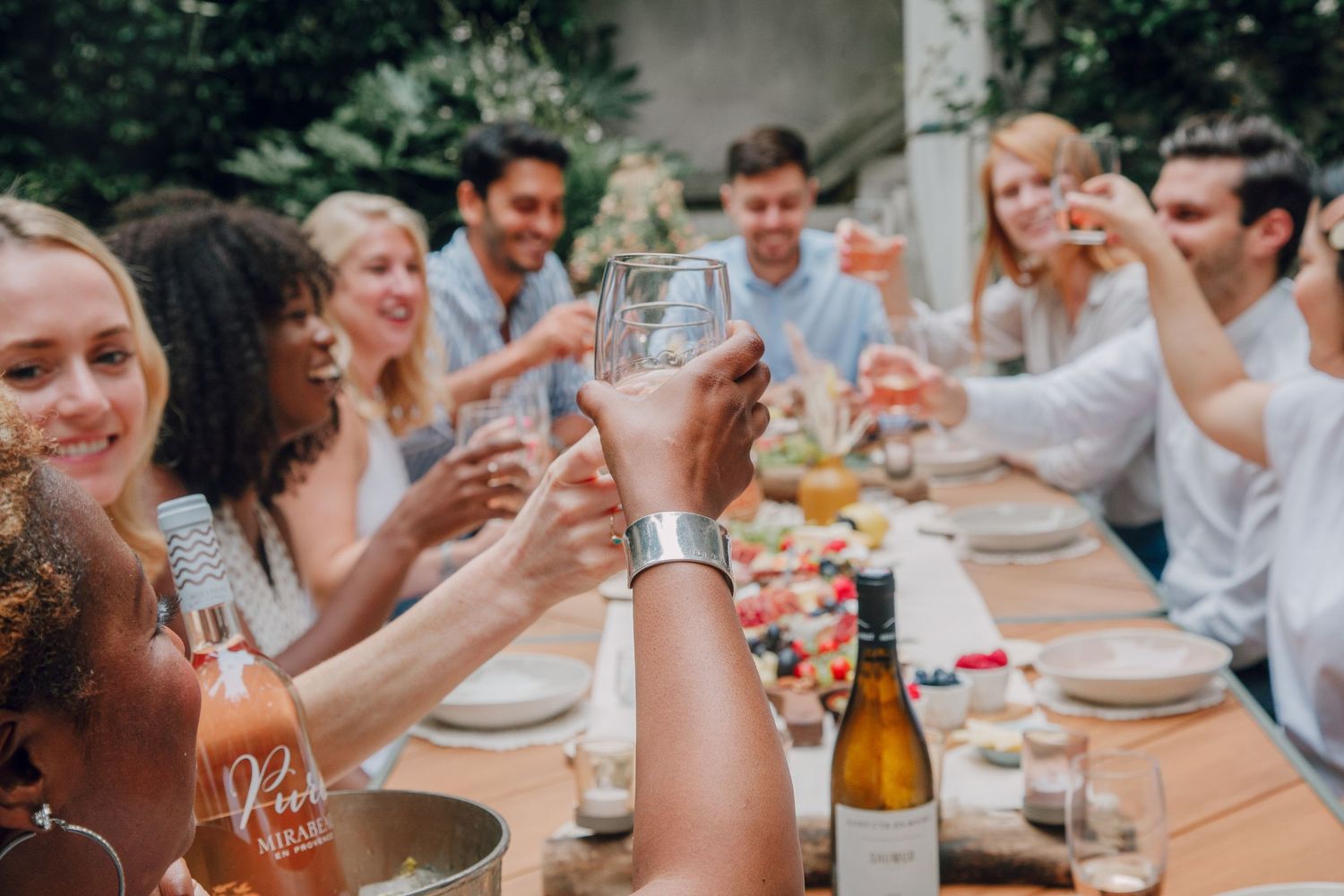 People enjoying a festive outdoor dinner party with drinks and laughter at a long table setting in natural light.