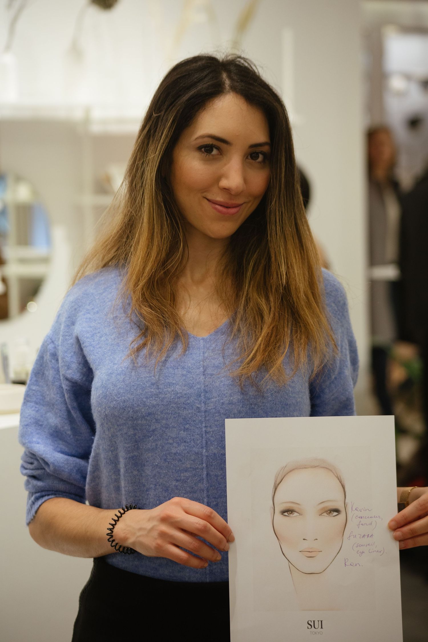 A person in a blue sweater holds up a simple face sketch showing makeup application techniques.