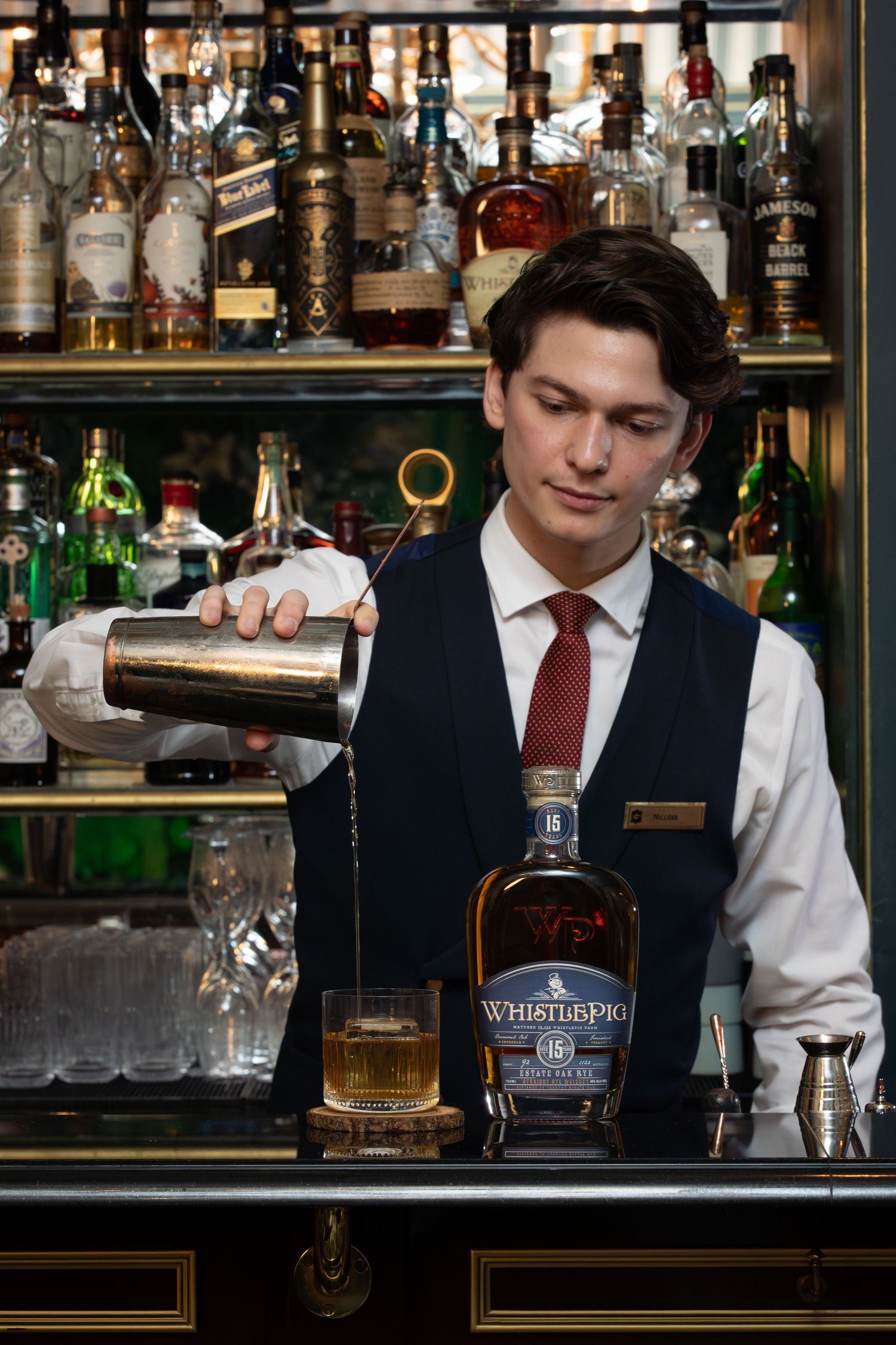 Professional bartender pours a whiskey drink at an elegant bar with bottles and glassware in the background.