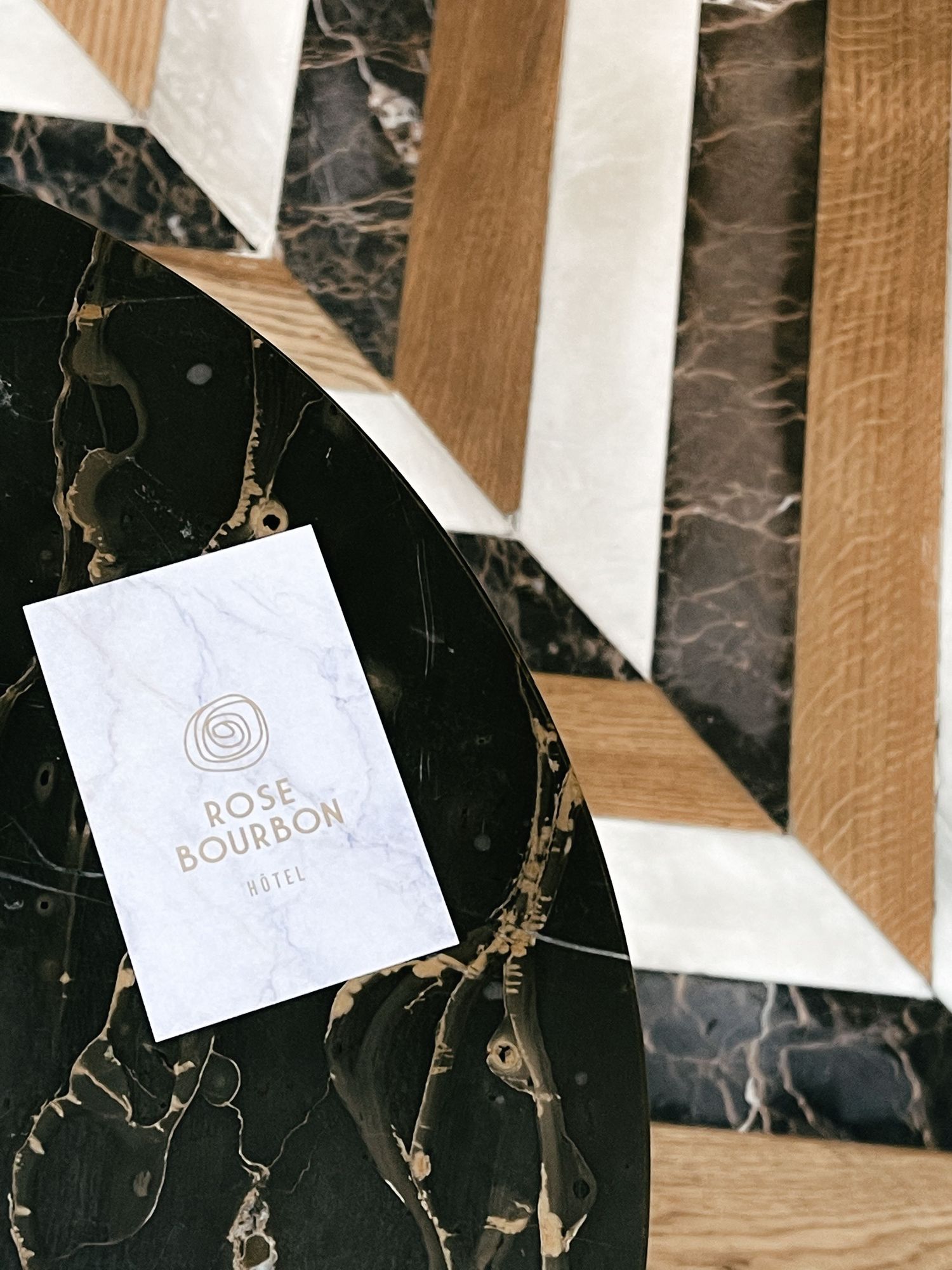 White business card with rose gold logo on black marble surface with wooden accents creating dramatic lighting and shadows.