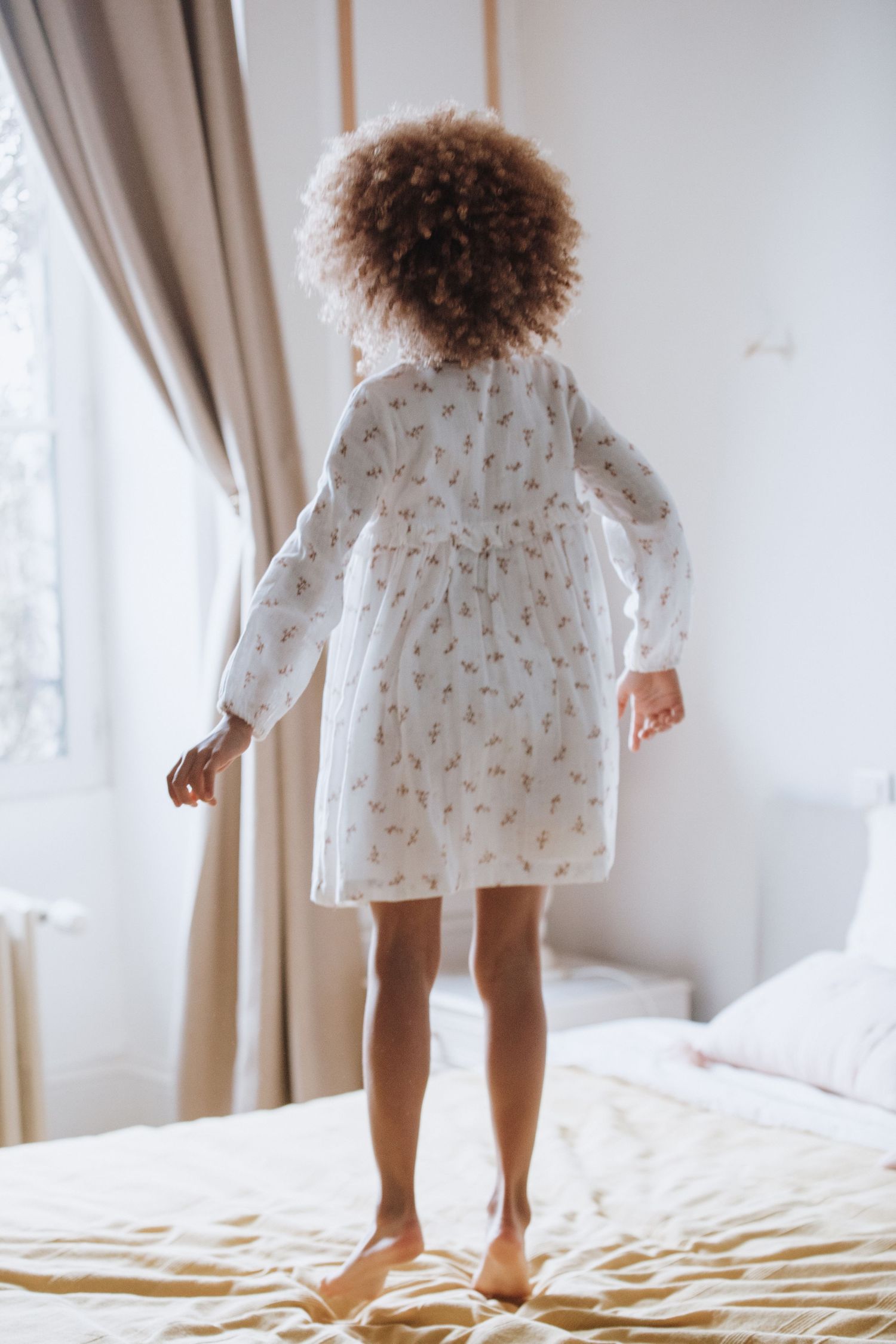 Back view of a child with curly hair wearing a white patterned nightgown while jumping on a bed near curtained windows.