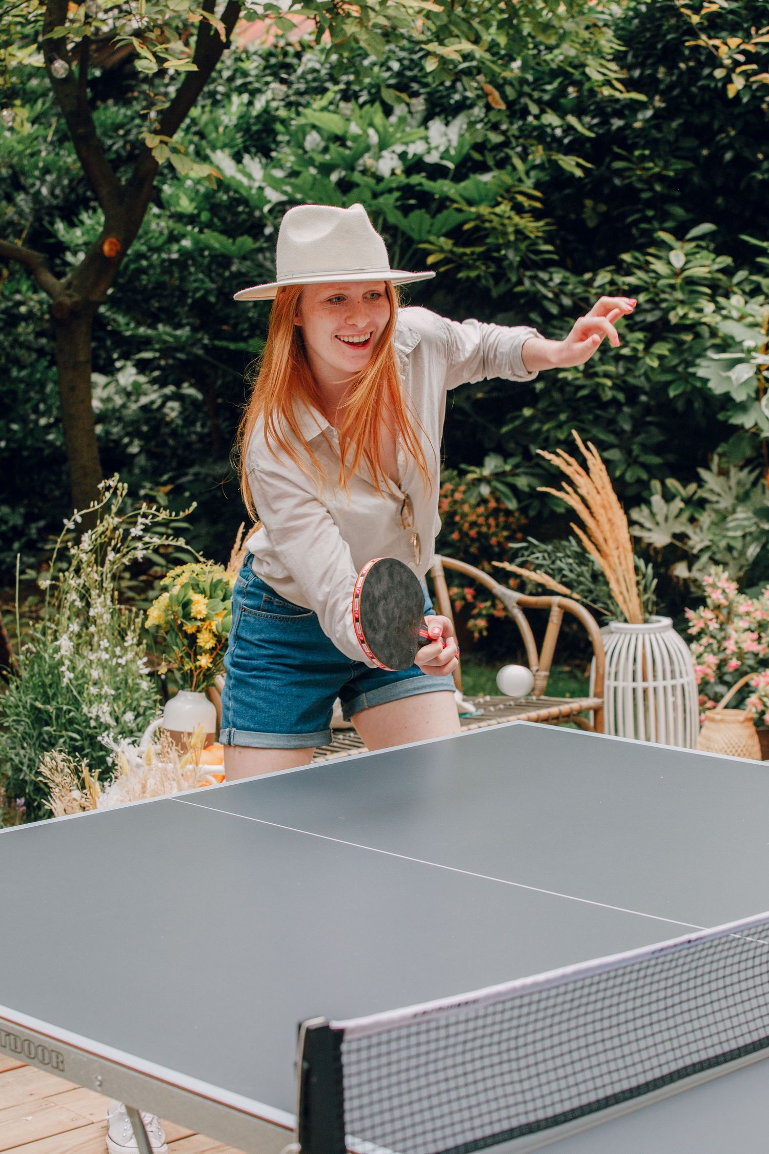 A person in a white sunhat and blue jeans plays ping pong outdoors on a summer day.