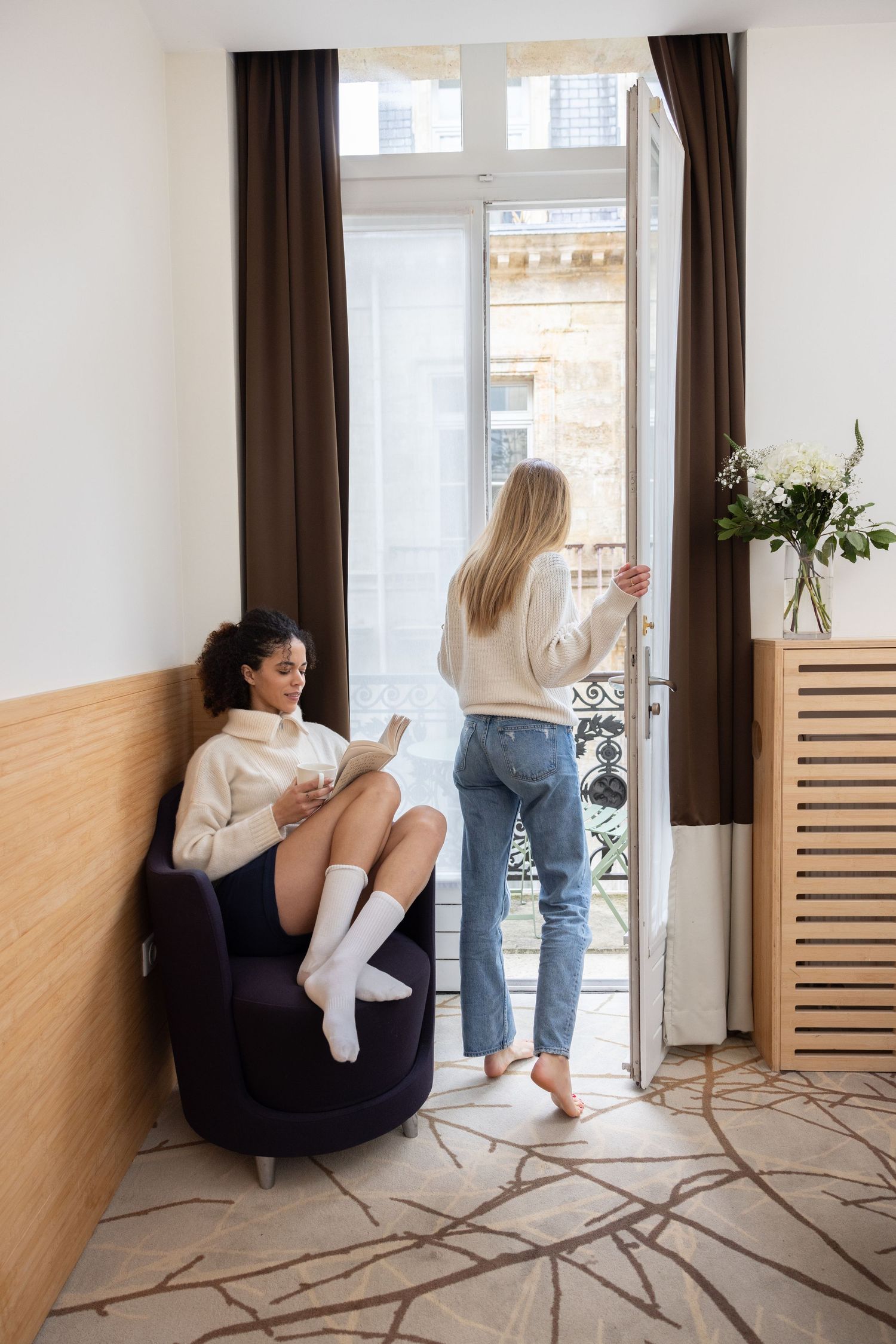 A person in jeans sits casually on a white floor against a wooden wall panel in a bright modern room.
