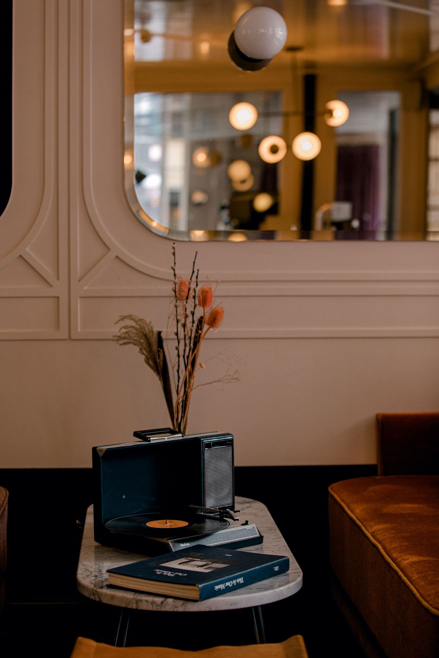 Cozy cafe interior with dried flowers in vase, vintage record player and books on marble table under warm lighting.