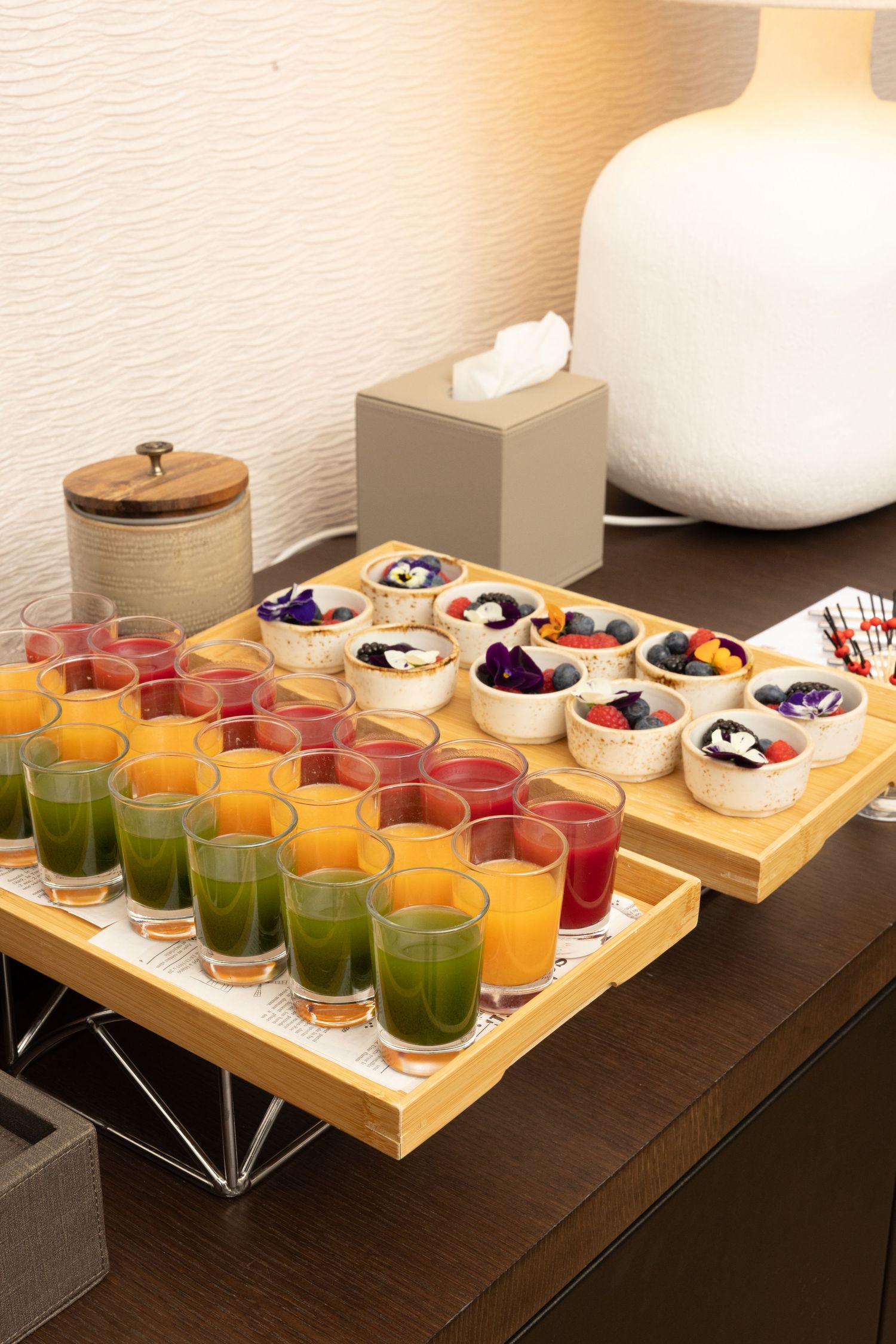 Wooden serving trays display colorful juice shots and yogurt parfaits with berries arranged in neat rows on a dark table.
