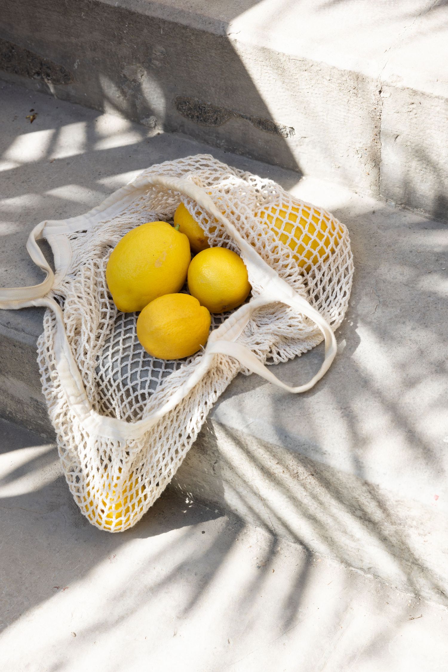 Fresh yellow lemons in a white mesh produce bag casting soft shadows on a light surface.