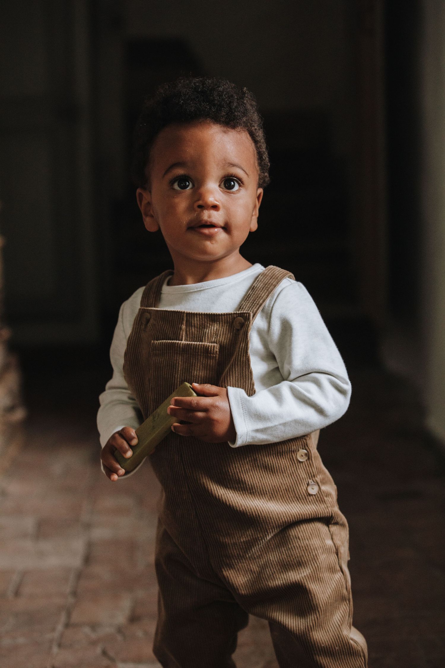 Small child wearing brown corduroy overalls and white long sleeve shirt stands in moody indoor lighting.