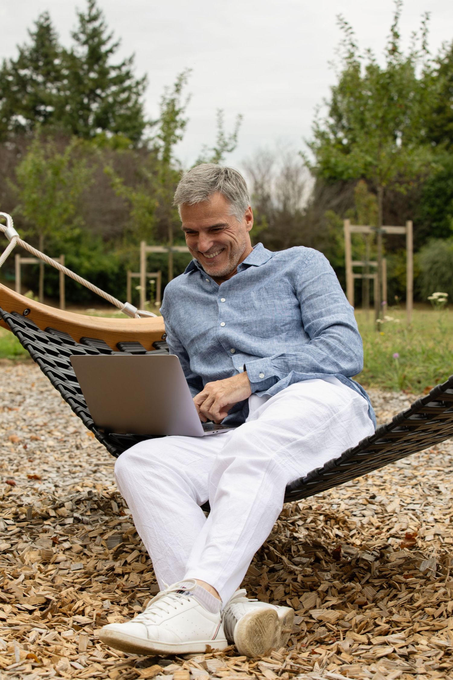 A person in white pants and blue shirt relaxes in a hammock while using a laptop in an outdoor garden setting.