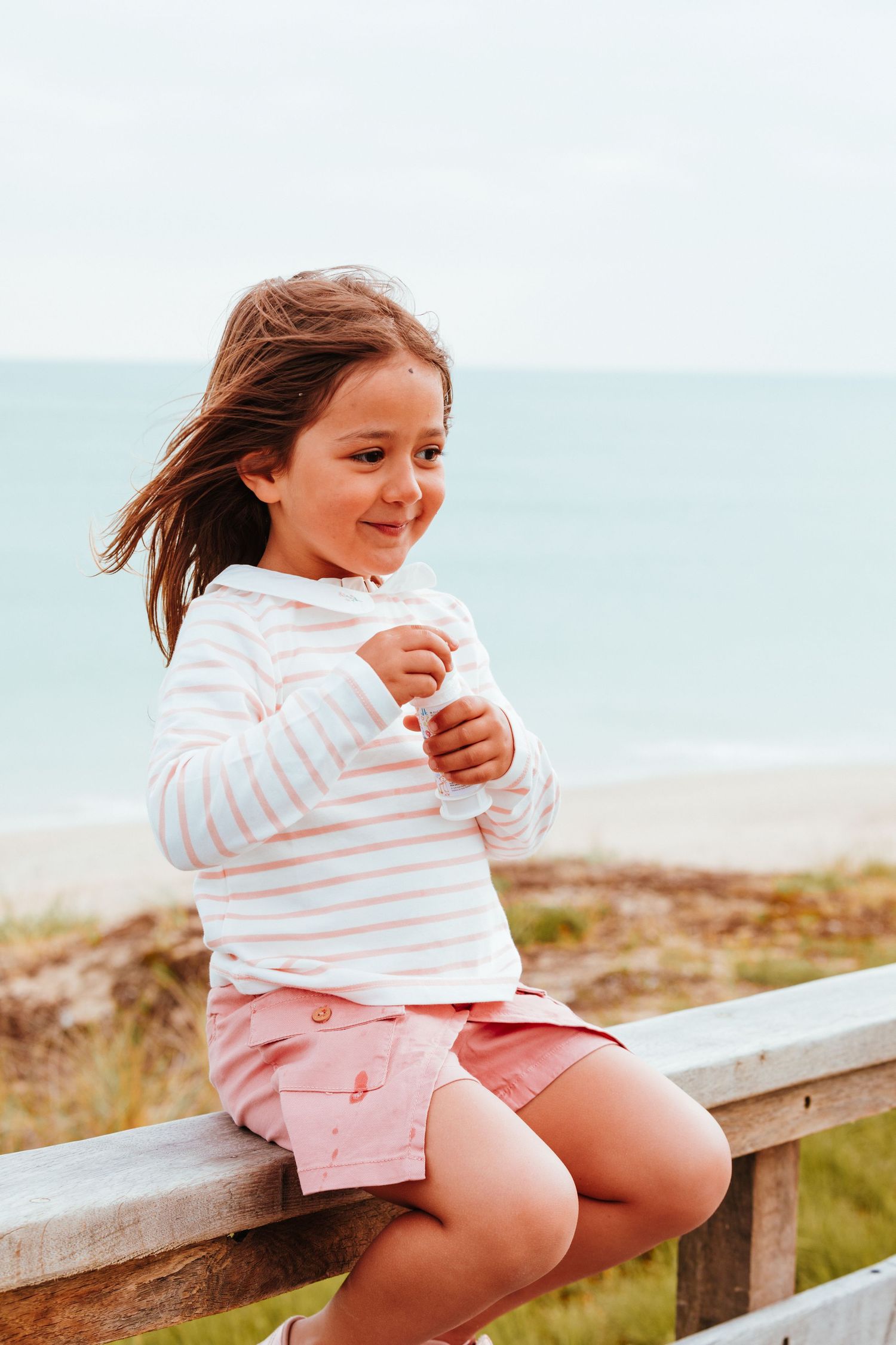 A child in a striped white sweater and pink shorts sits on a wooden railing at the beach with windswept hair.