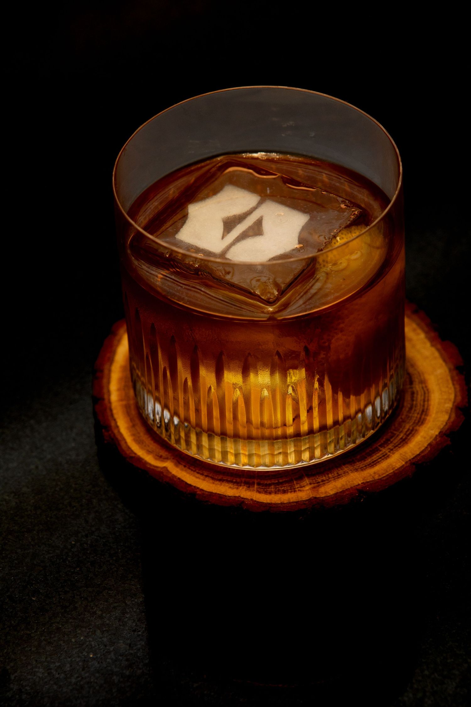 A glowing amber cocktail with ice cubes served in an elegant glass on a dark surface creates a dramatic lighting effect.