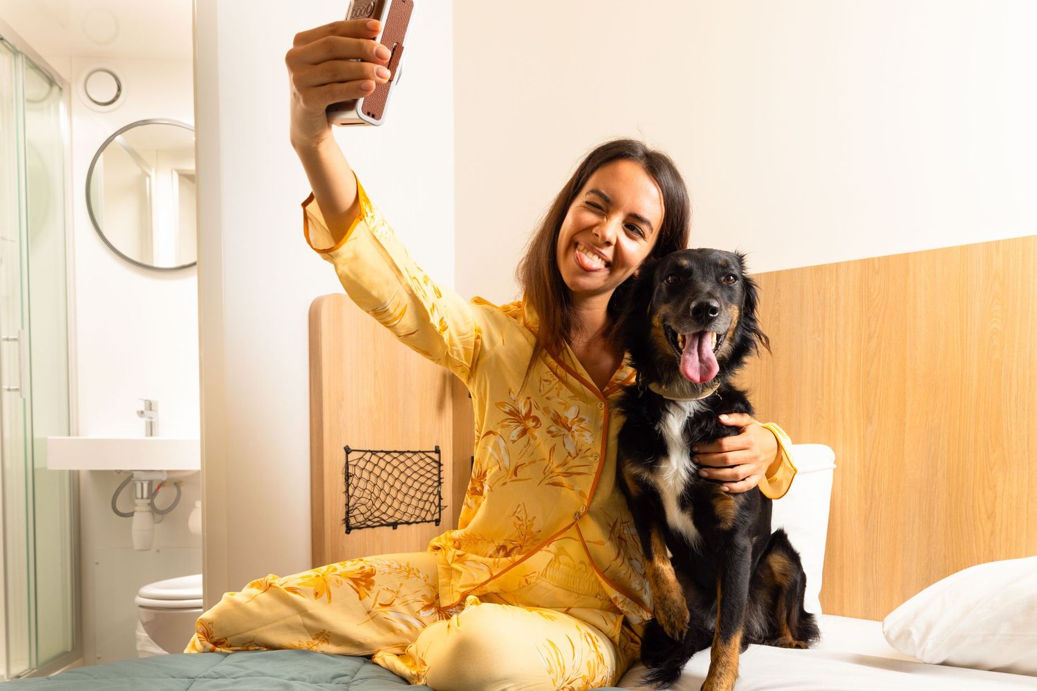 Someone in a yellow dress takes a selfie while sitting on a bed with a black dog in a bright interior setting.