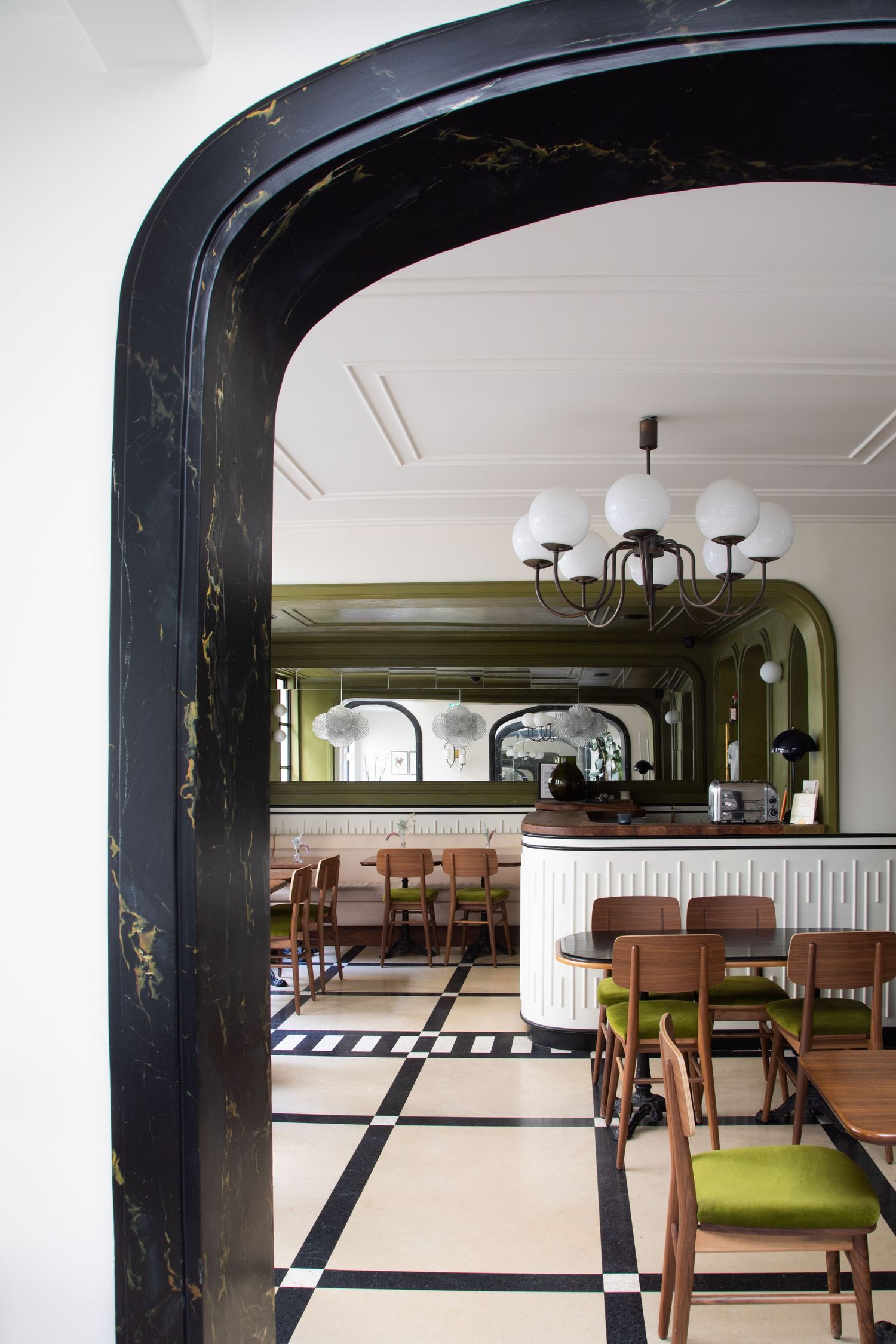 A restaurant interior with checkered floor tiles, wooden tables, and green upholstered seating visible through an archway.