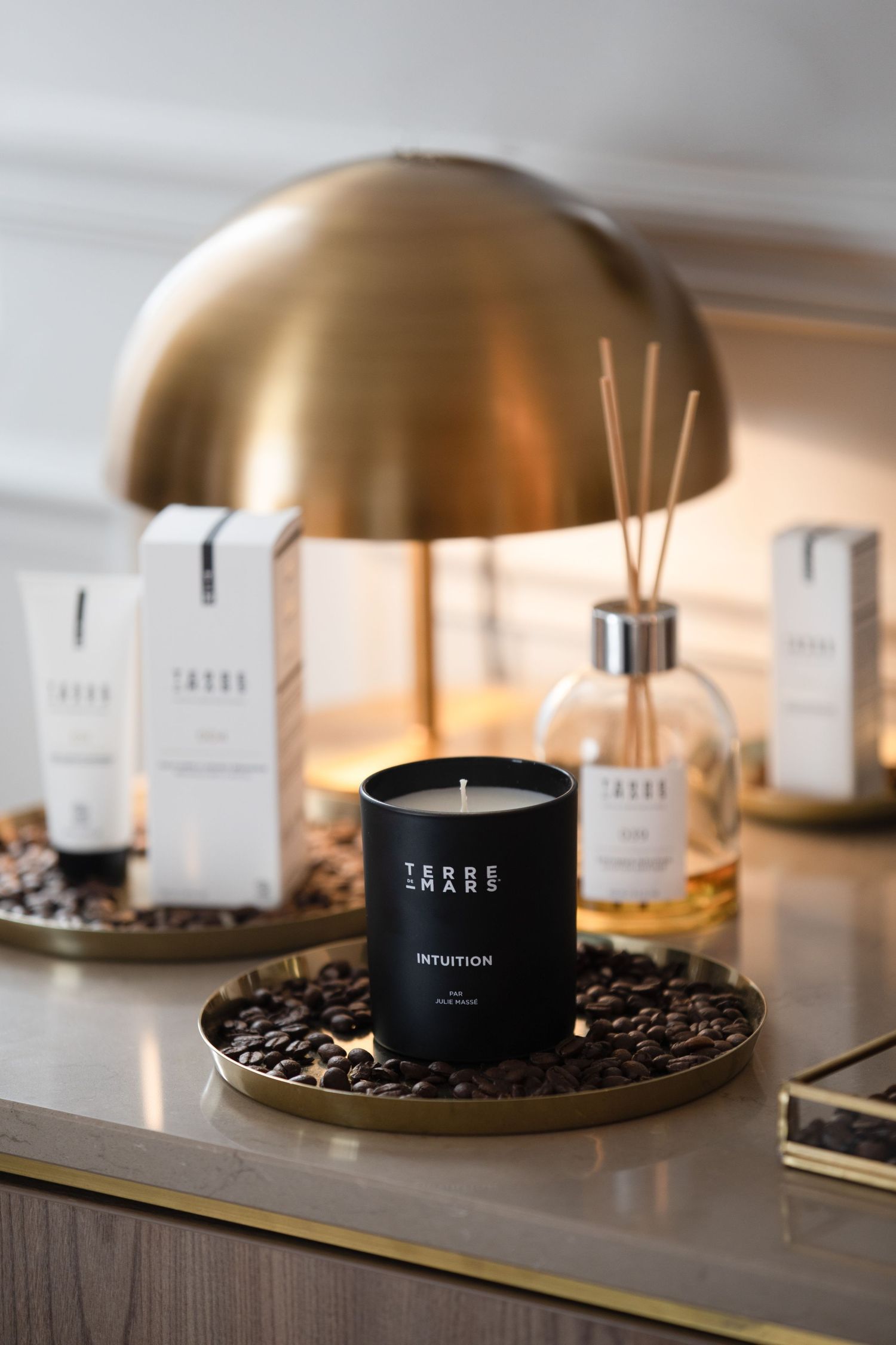 A series of minimalist black candles and reed diffusers arranged on wooden surfaces with brass accents and coffee beans.