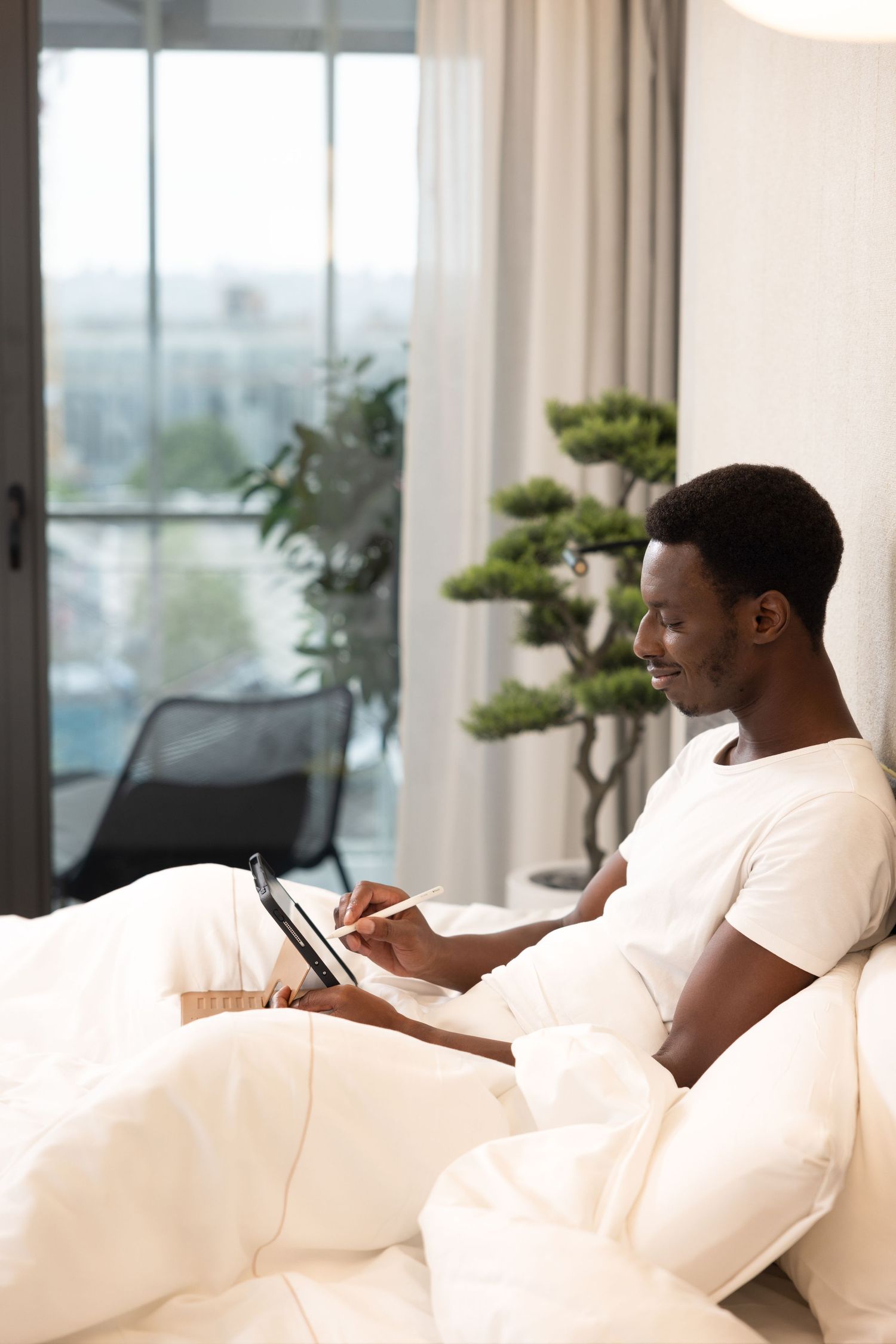 Person relaxing on white couch using mobile phone in modern apartment with large windows and natural light.