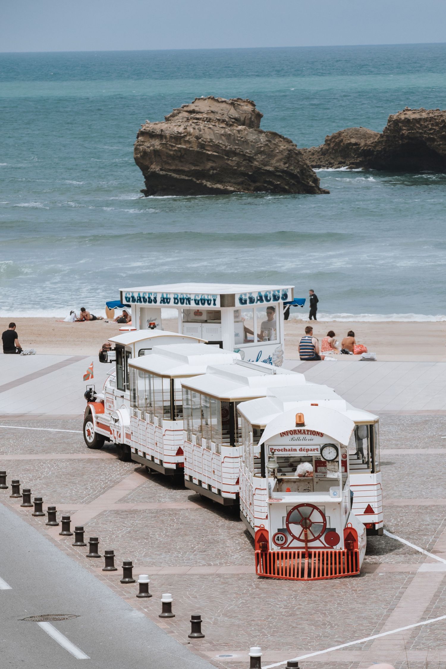 A white tourist train parked on a beach promenade with rocky cliffs and turquoise ocean waters in the background.