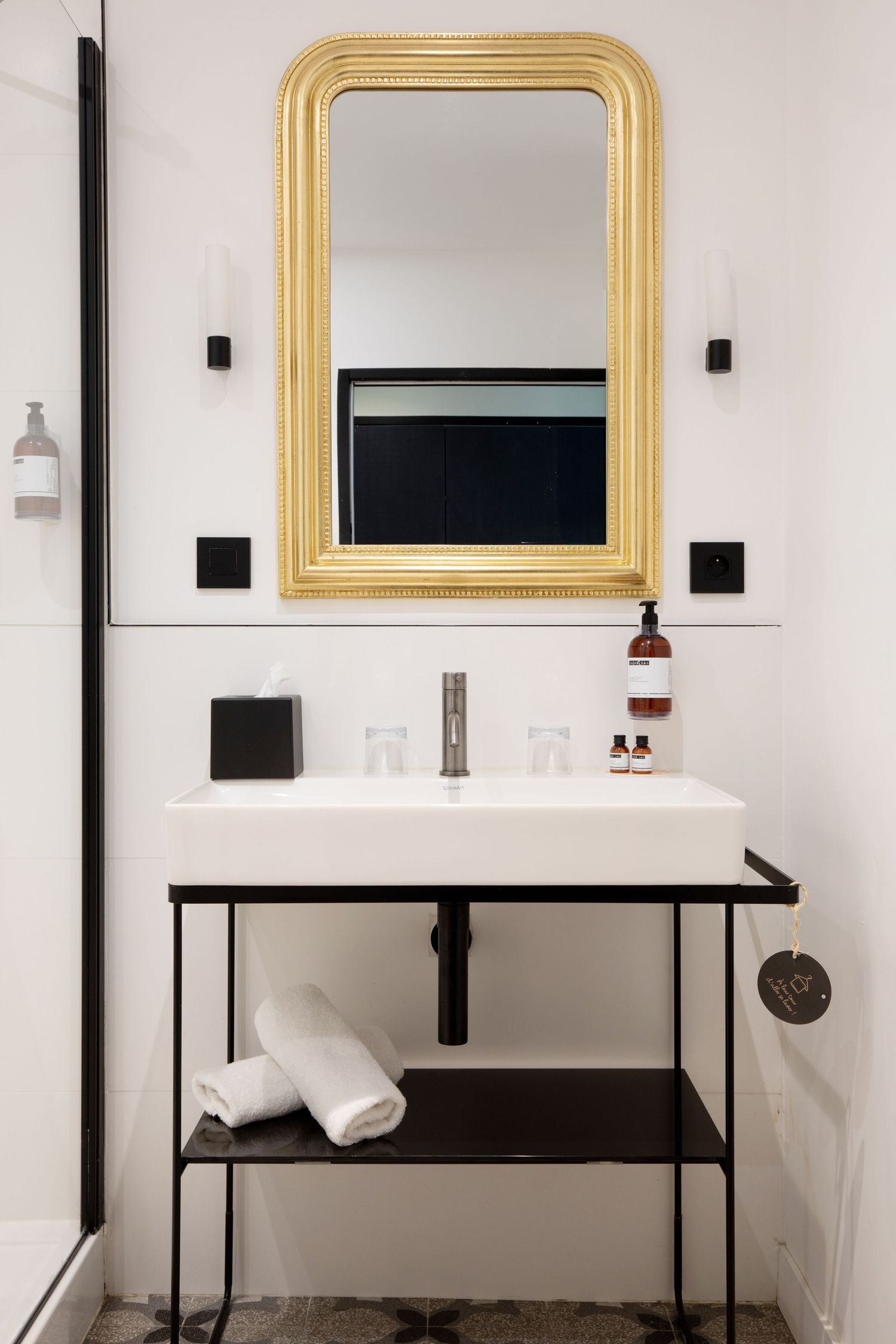 Minimalist bathroom vanity with black metal frame, white sink, gold-framed mirror and modern wall sconces.