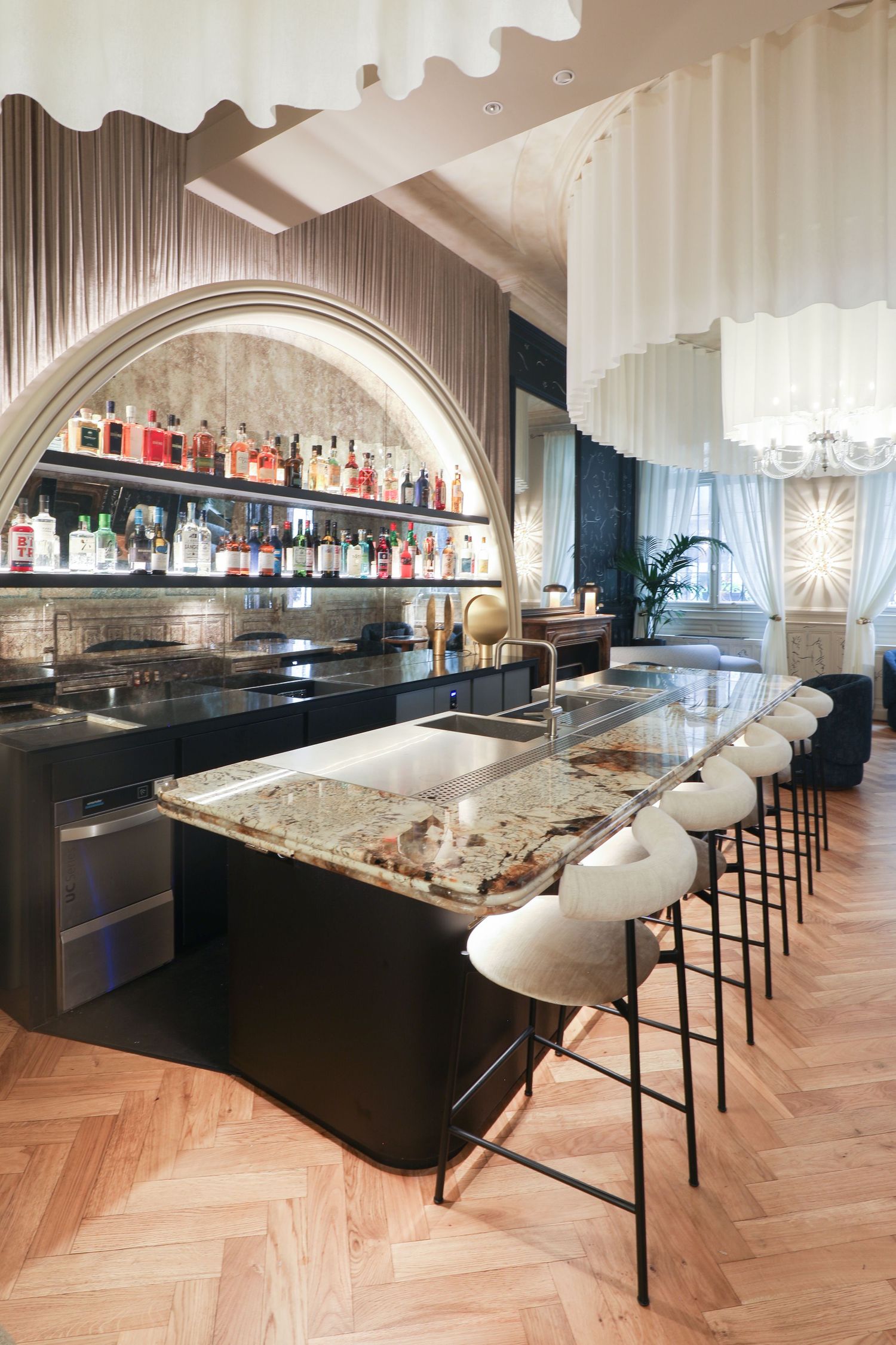 Modern luxury bar interior with marble countertop, white bar stools, and arched liquor display shelf on herringbone wood floors.