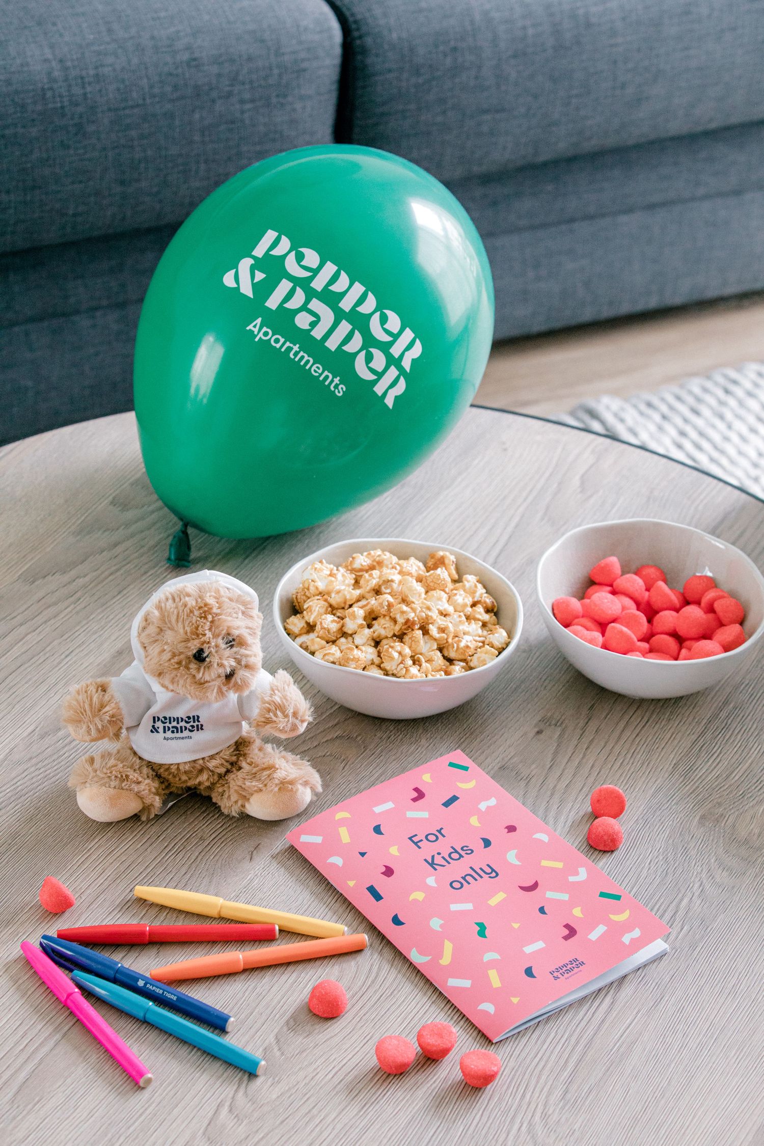 Flatlay of party supplies including green balloon, teddy bear, popcorn, pink candies, colored markers and pink notebook.