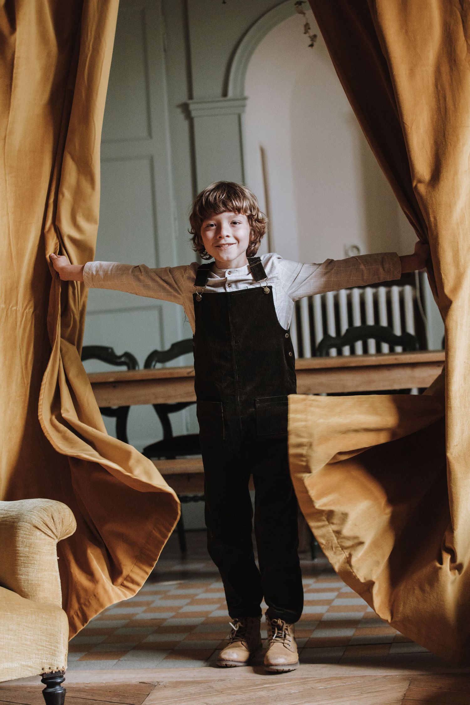 Small child in black clothing stands between flowing gold curtains in an elegant interior space.