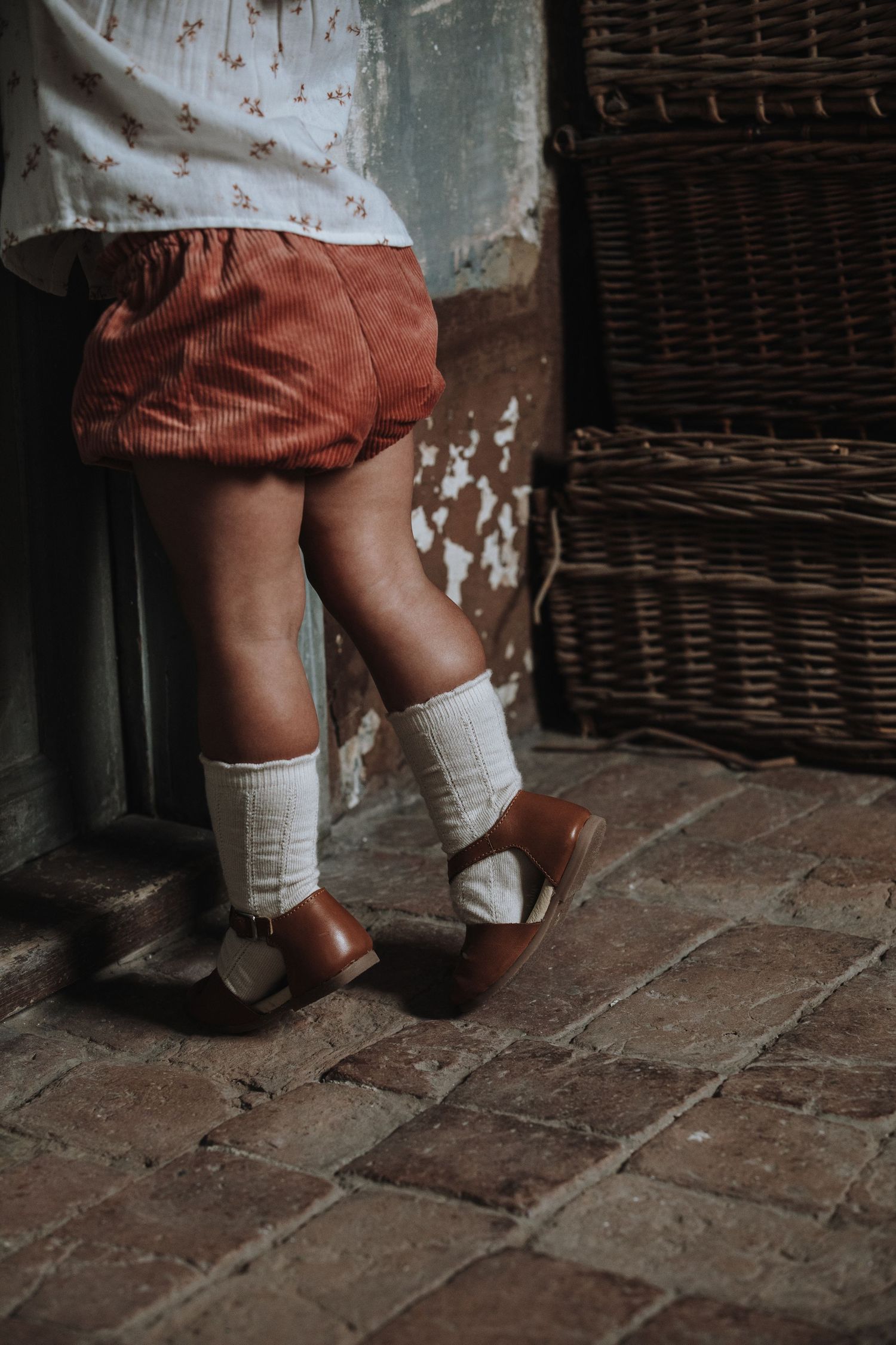 Small legs in corduroy shorts and white socks with brown leather shoes stand on rustic brick flooring near wicker furniture.