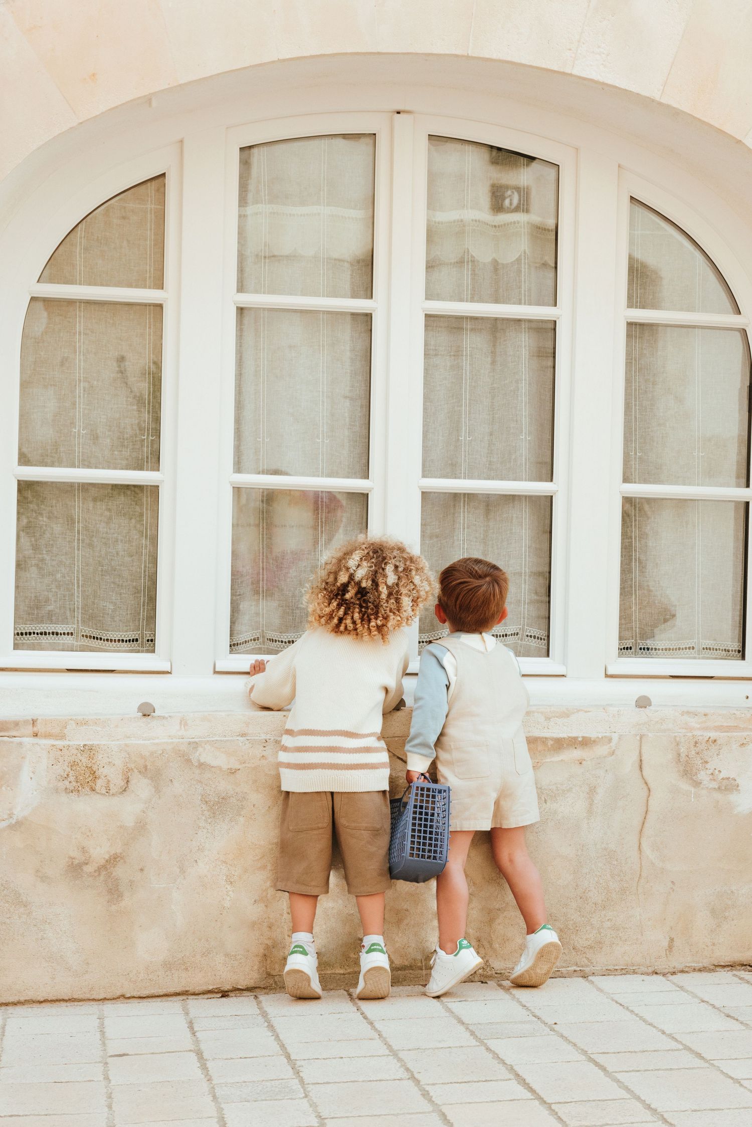 Two small children stand on tiptoes looking through a large arched window with white frames.
