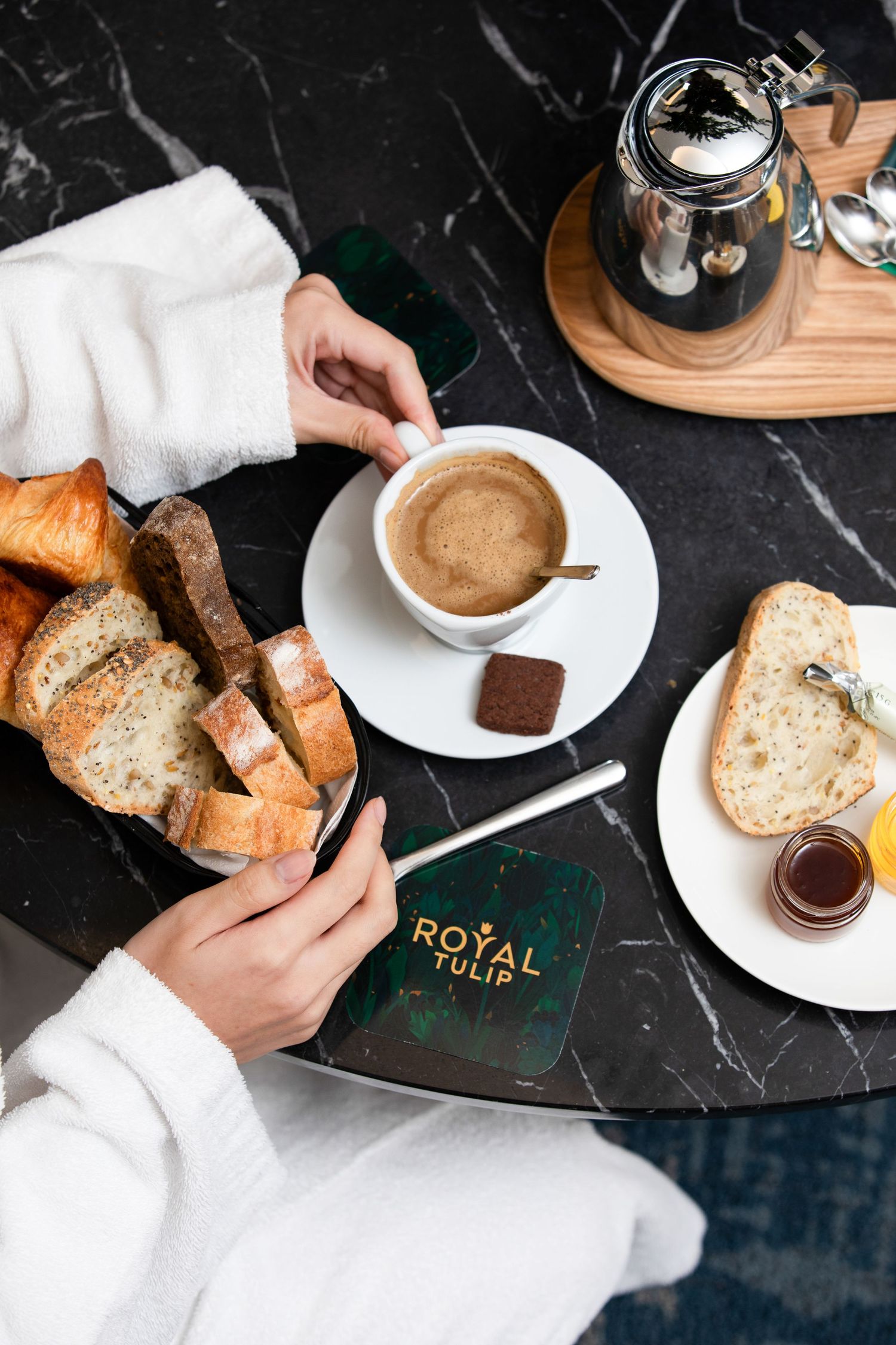 Hands cutting rustic bread slices on dark surface with coffee cup and chocolate spread jar in moody lighting setup.