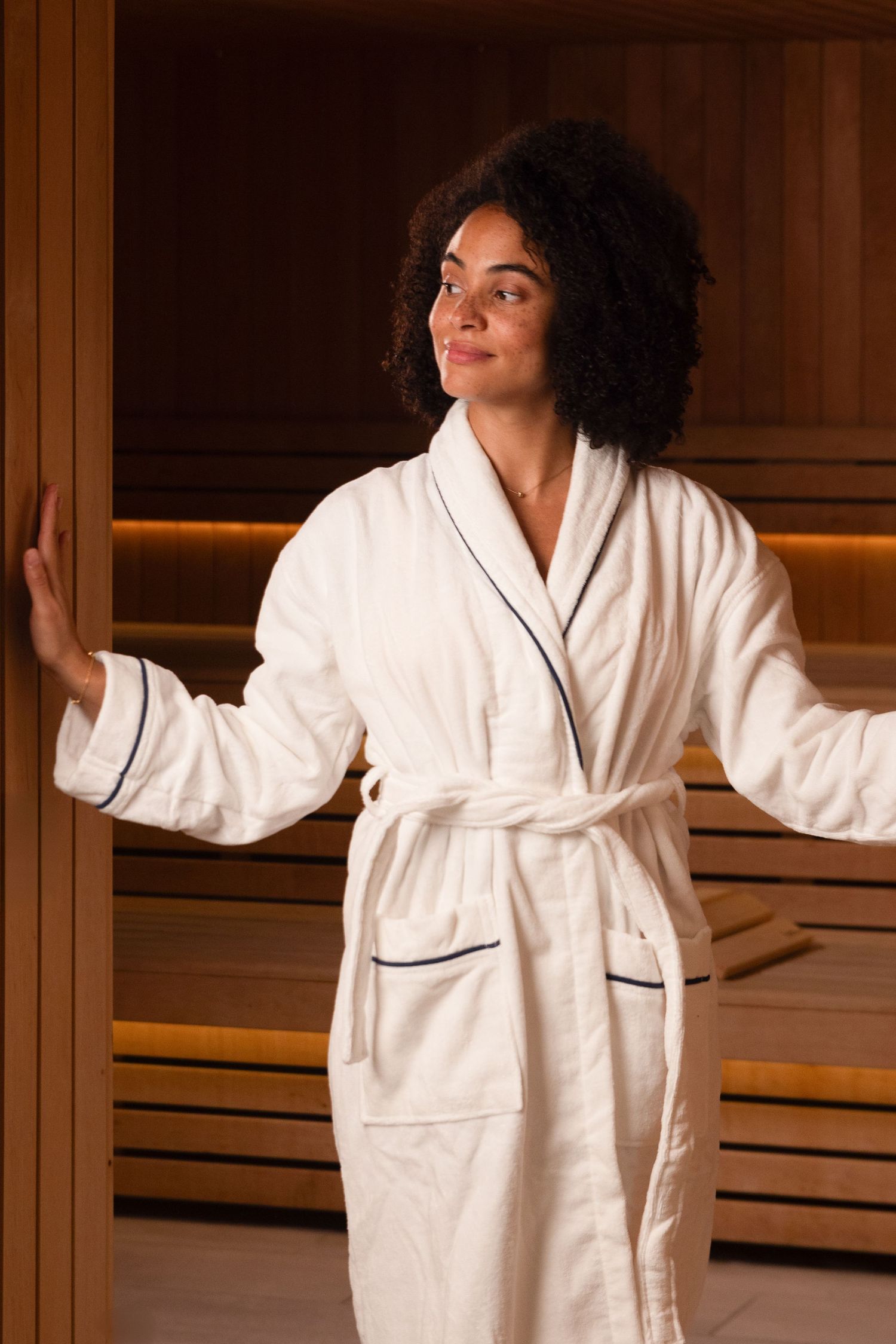 Person wearing white bathrobe relaxing in wooden sauna or spa room with warm lighting.