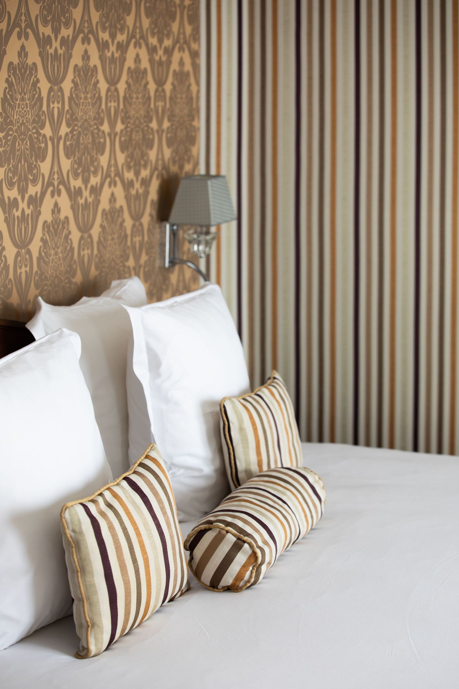 Elegant bedroom interior with striped wallpaper and white bedding accented by brown and beige striped throw pillows.